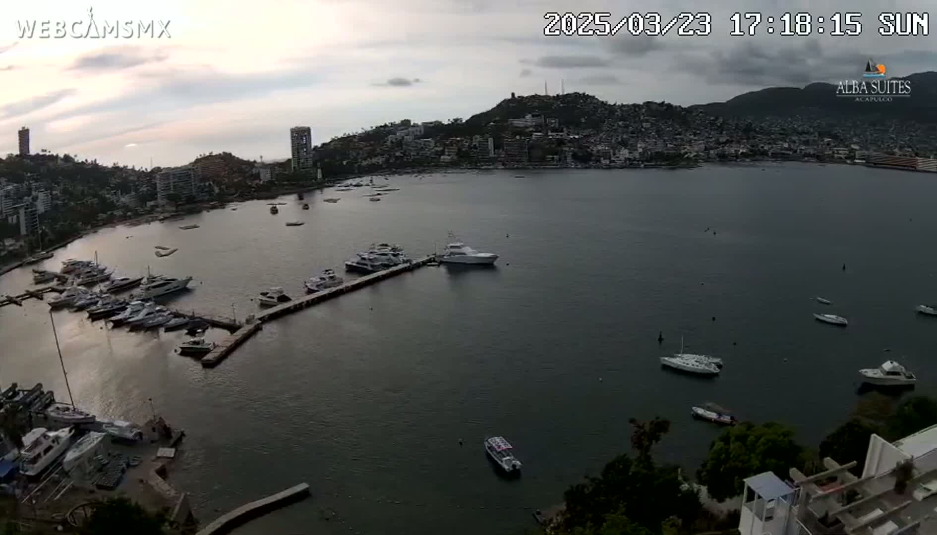 A calm bay with numerous boats docked at a marina, situated in front of a city built on a hillside under a partly cloudy sky.