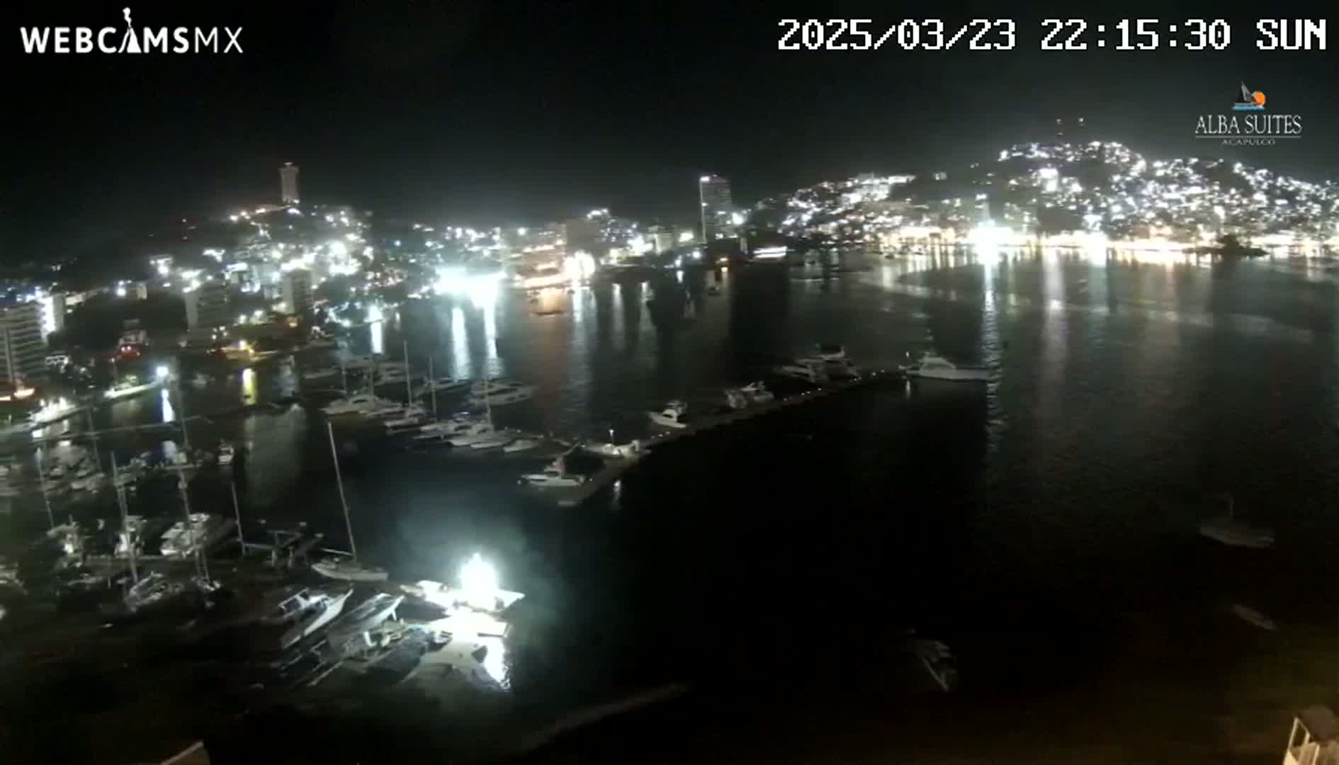 A nighttime view of a harbor filled with boats, next to a brightly lit city on a calm, dark water.