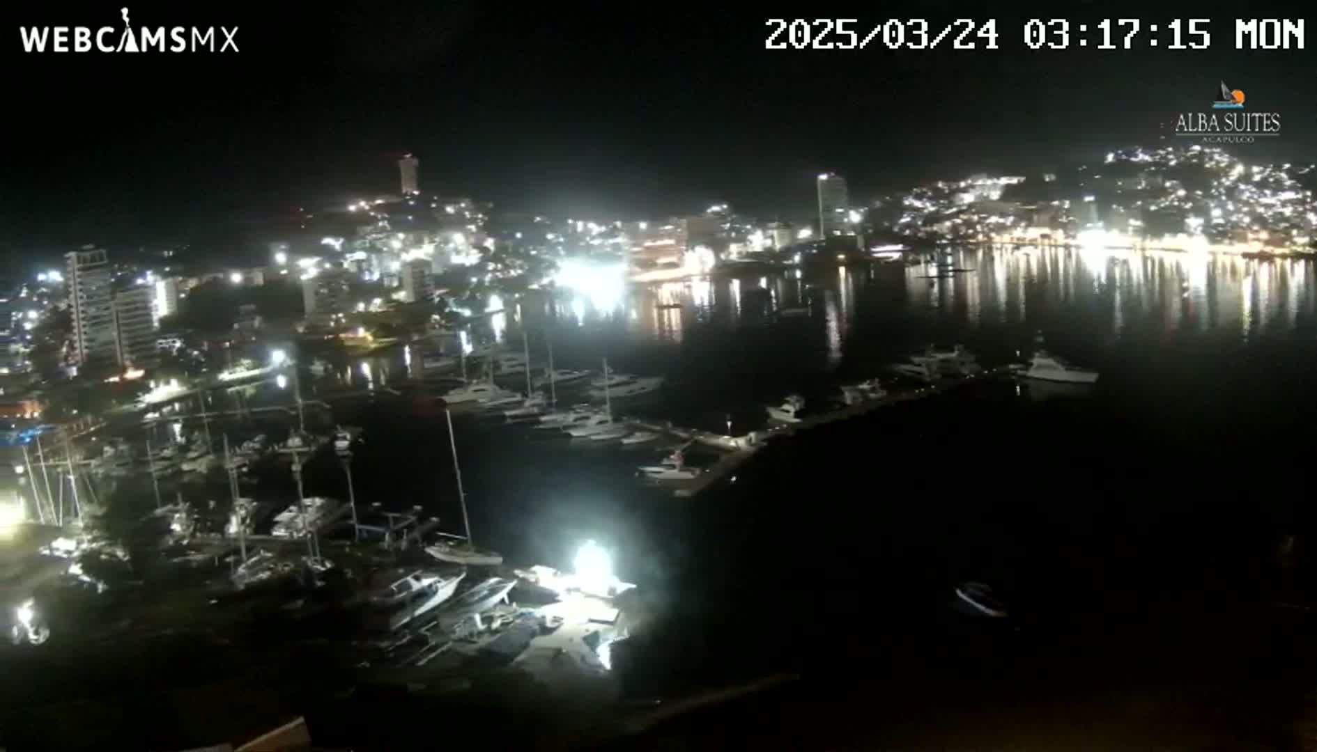 A nighttime view of a city harbor filled with boats, illuminated by city lights reflecting on calm water.