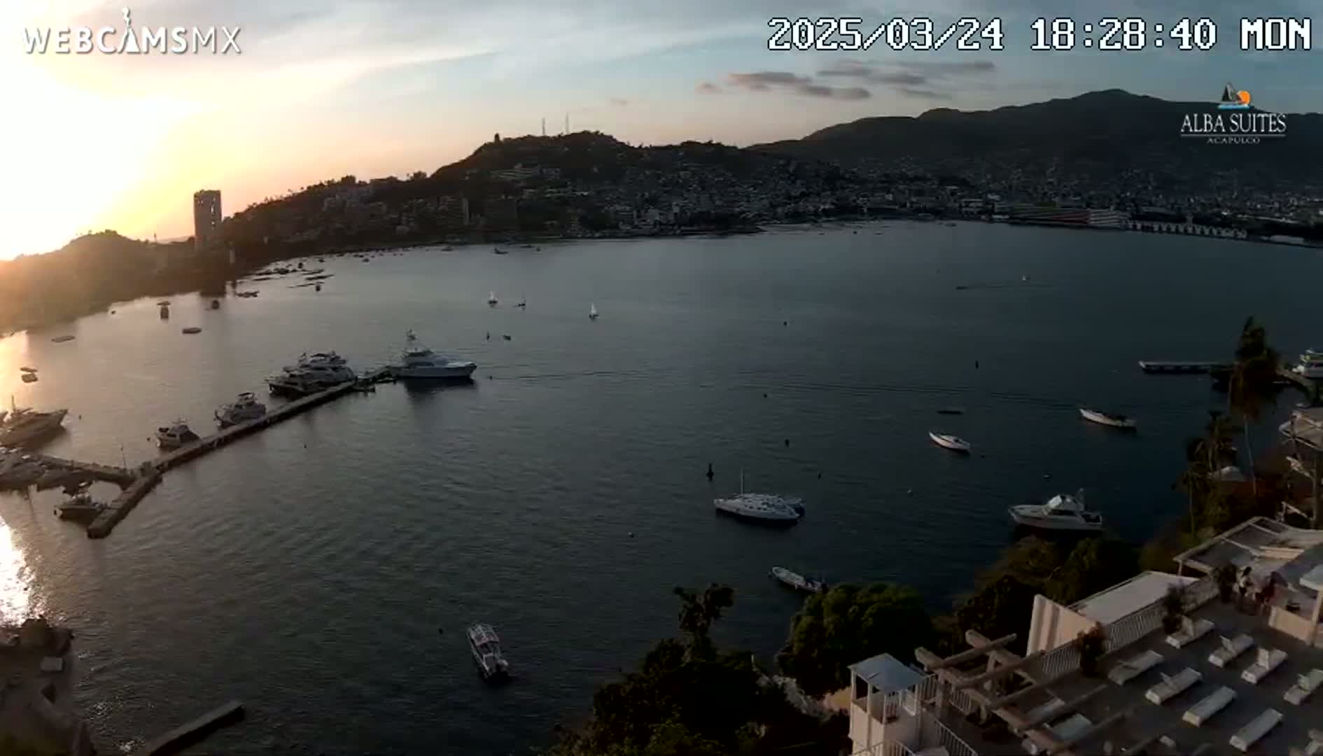 A sunset view over a calm bay with numerous boats docked and anchored, hills in the background, and a partially visible building with lounge chairs in the foreground.