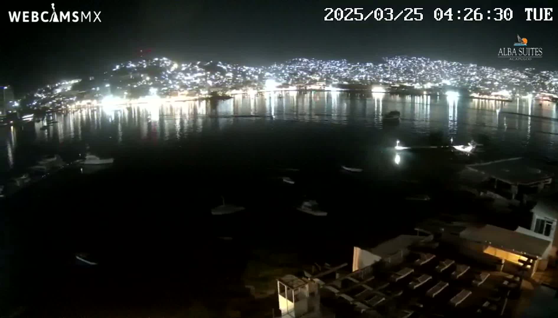 A nighttime view of a brightly lit city across a dark body of water with several boats, calm water reflecting the city lights.