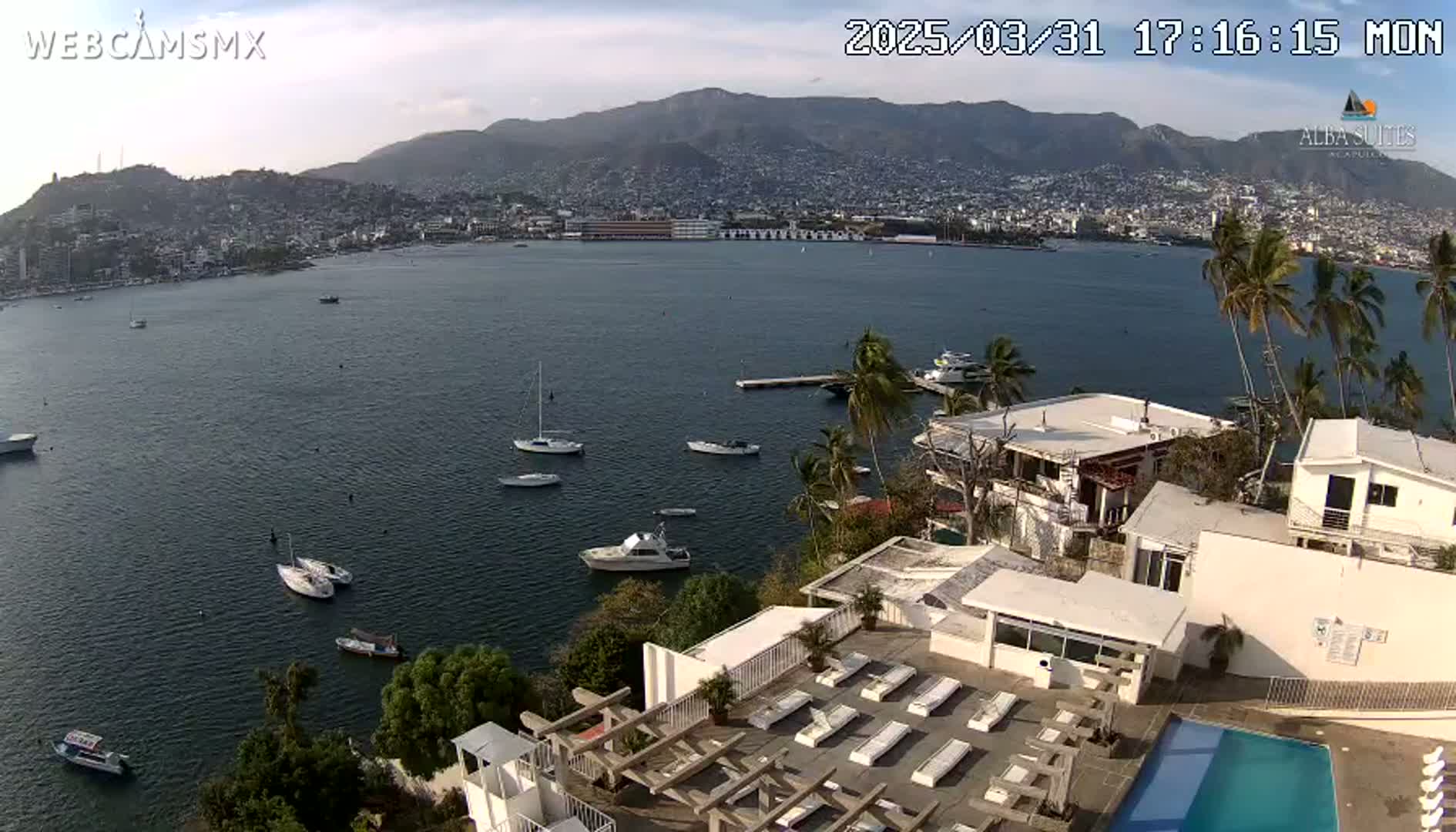 An aerial view shows a calm bay with several boats, a city nestled in mountains in the background, and a white building with a swimming pool and sun loungers in the foreground, under mostly clear skies.