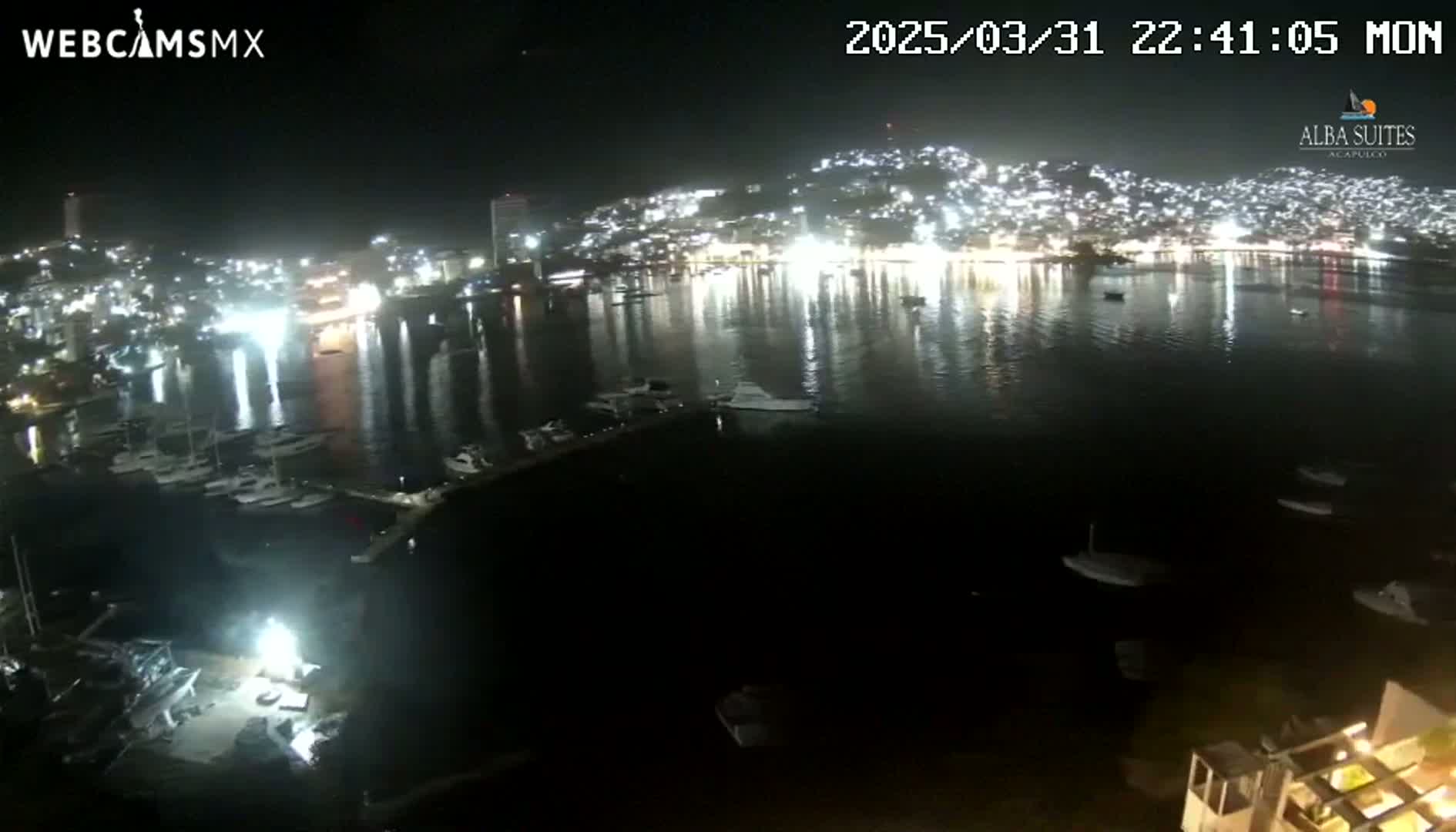 A nighttime view of a harbor filled with boats, reflecting the lights of a brightly lit city on a hill across the water under a clear sky.