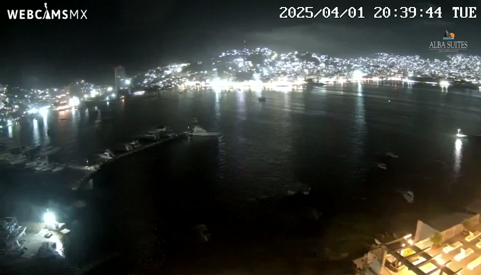 A nighttime, calm-water view of a brightly lit city across a bay, with boats moored in the foreground.