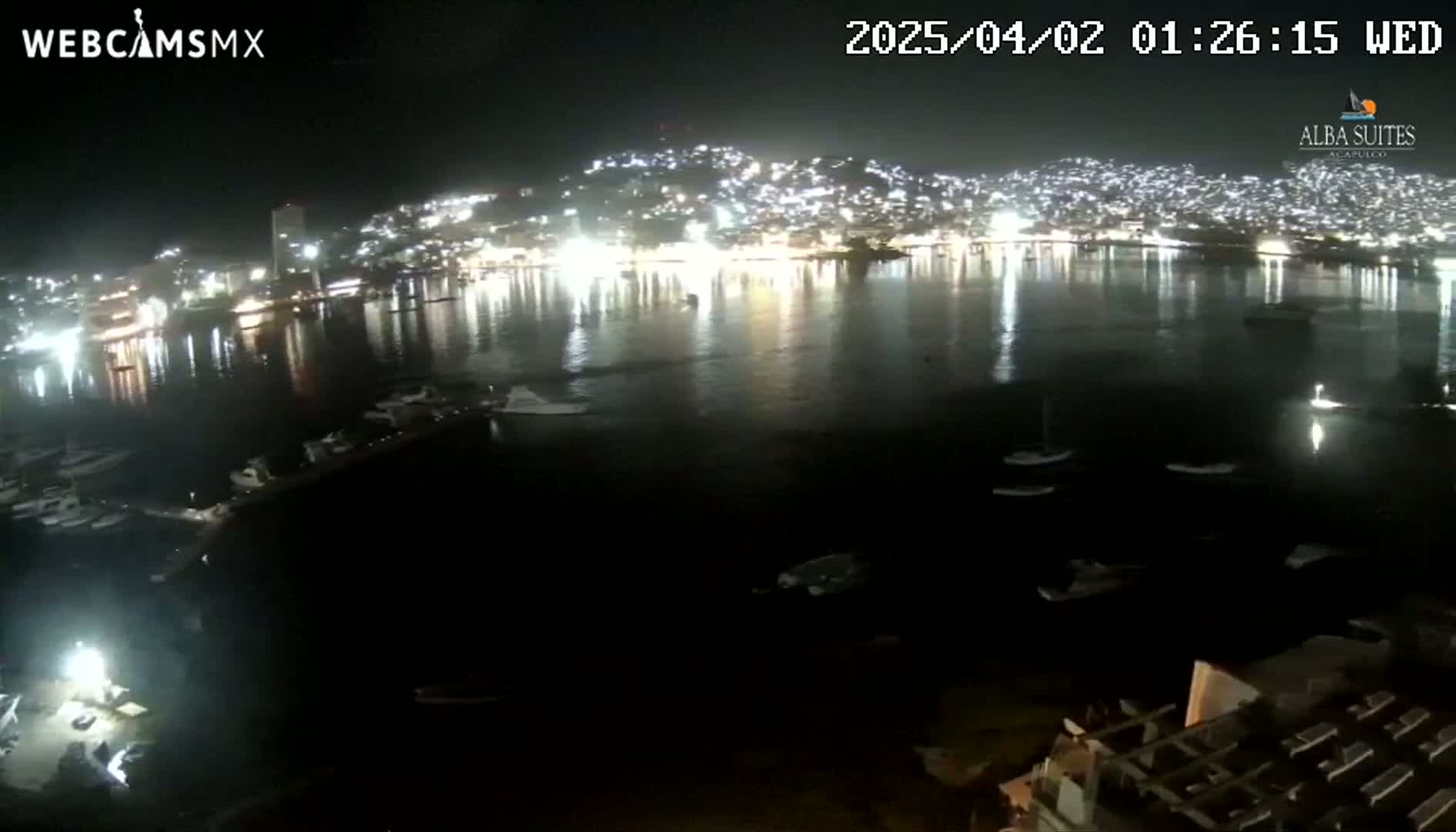 A nighttime view of a brightly lit city across a dark body of water where several boats are moored.