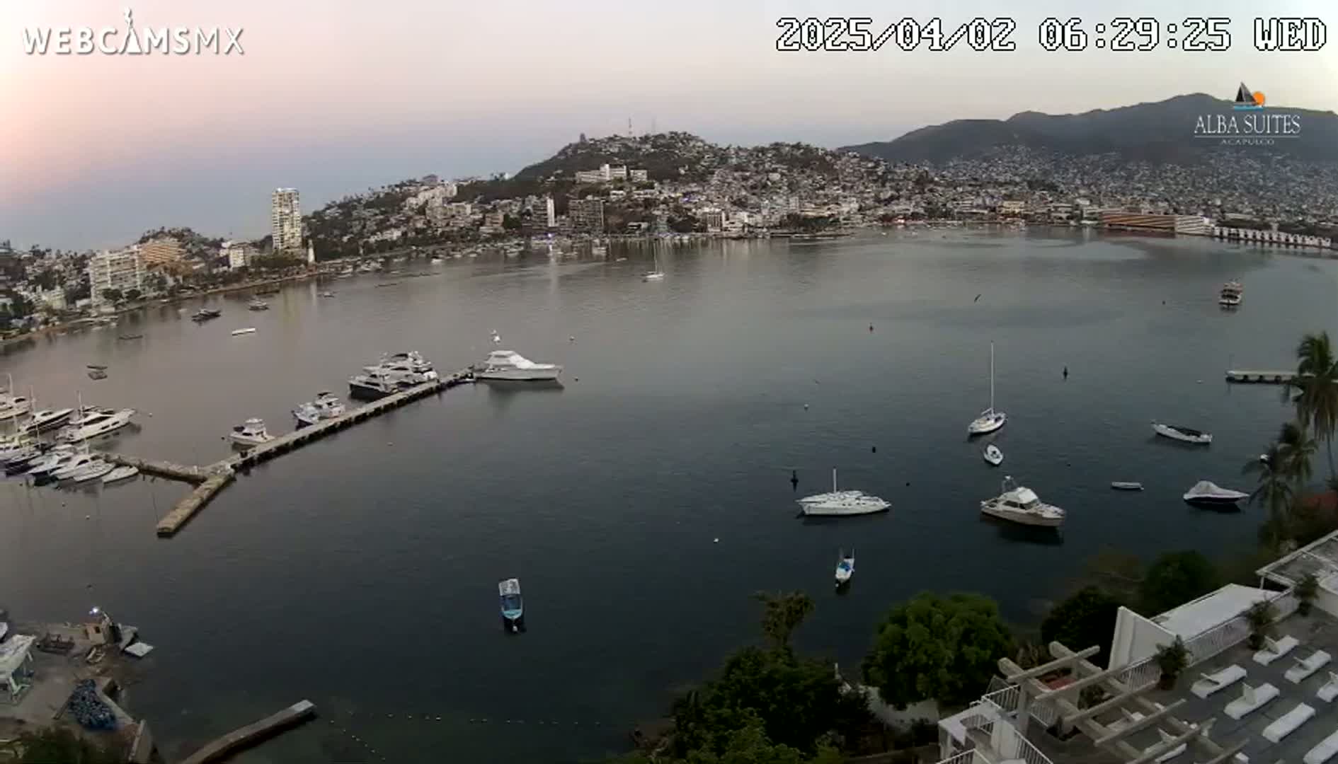 A calm bay with numerous boats docked at a marina, nestled beside a hillside town under a twilight sky.
