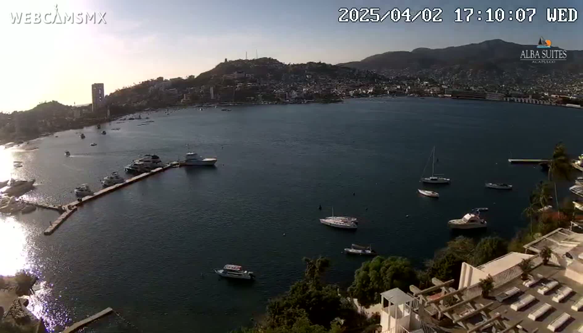 A high-angle, long shot shows a calm bay with numerous boats and yachts docked near a city nestled against hills under a clear, sunny sky.