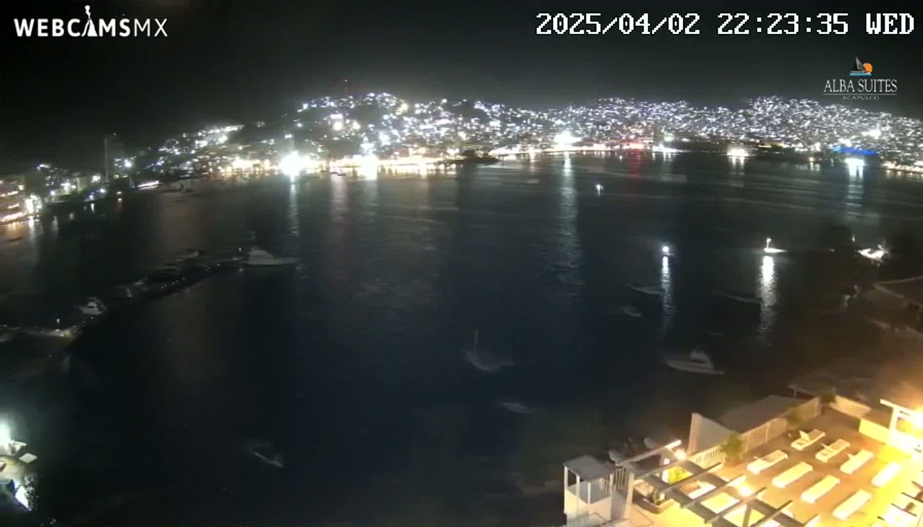 A nighttime view of a brightly lit city across a calm bay, with several boats visible in the water and a lighted patio area in the foreground.