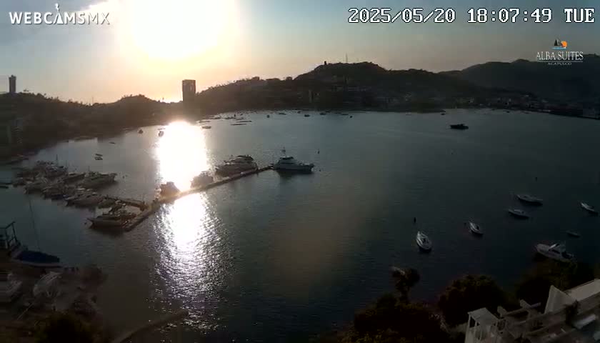 A tranquil harbor scene at sunset, featuring numerous boats and yachts in a calm bay, with hills and buildings visible in the background.