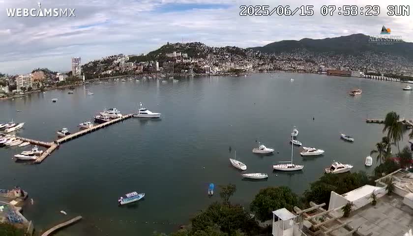 A calm bay with numerous boats and yachts docked at a pier, overlooking a city nestled against a hillside under a partly cloudy sky.