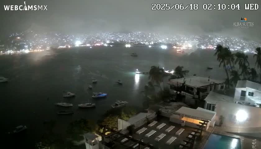A nighttime view of a rain-swept city and harbor, with numerous boats and a resort with a pool visible in the foreground.
