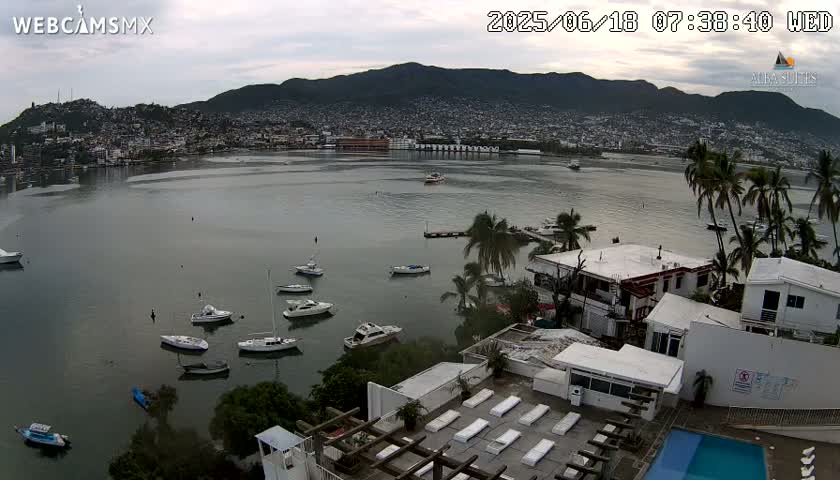 An overcast day reveals a calm bay filled with numerous boats, a city nestled against a mountain range in the background, and a rooftop pool area with lounge chairs in the foreground.