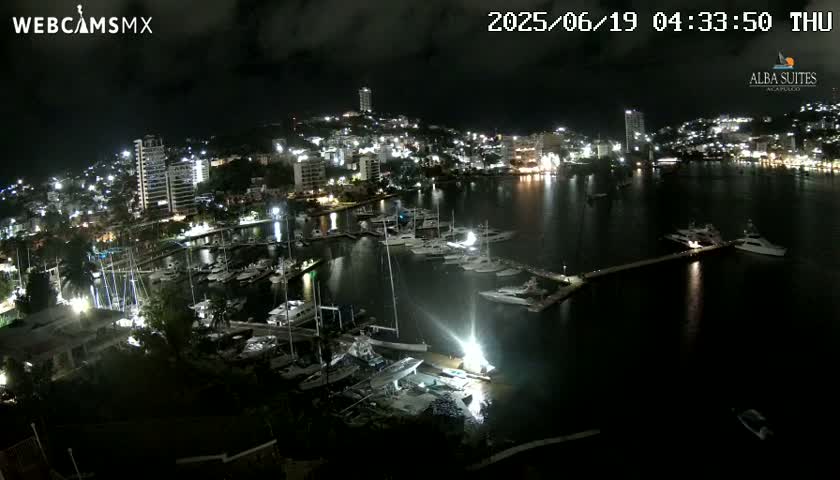 A nighttime aerial view shows a marina filled with boats, next to a city illuminated with lights under a dark sky.