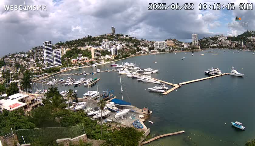 A marina filled with yachts and boats is situated in a bay next to a city on a partly cloudy day.