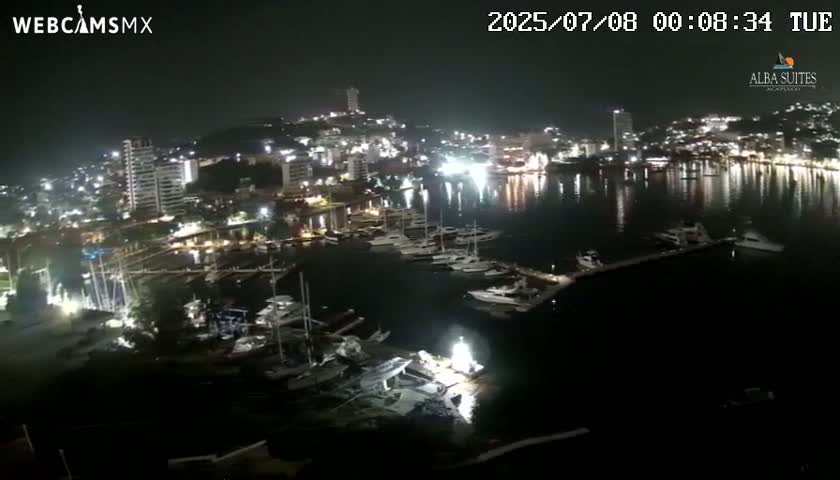 A nighttime, brightly lit cityscape with a harbor full of boats is reflected in calm water under a clear sky.