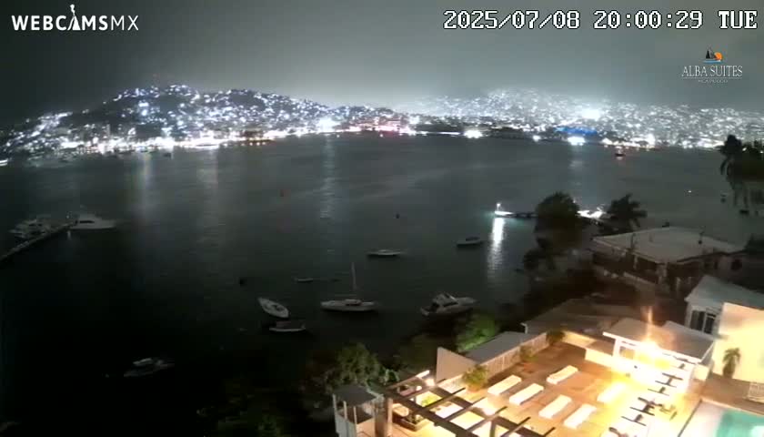 A nighttime view of a brightly lit city across a dark bay, with several boats on the water and a partially visible pool area at the bottom.