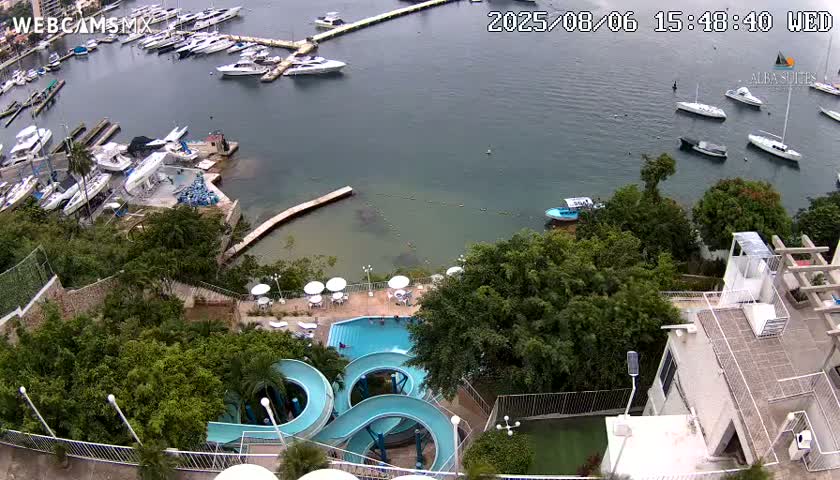 An overhead view shows a marina with numerous boats, a pool with water slides and surrounding patio furniture, all under an overcast sky.