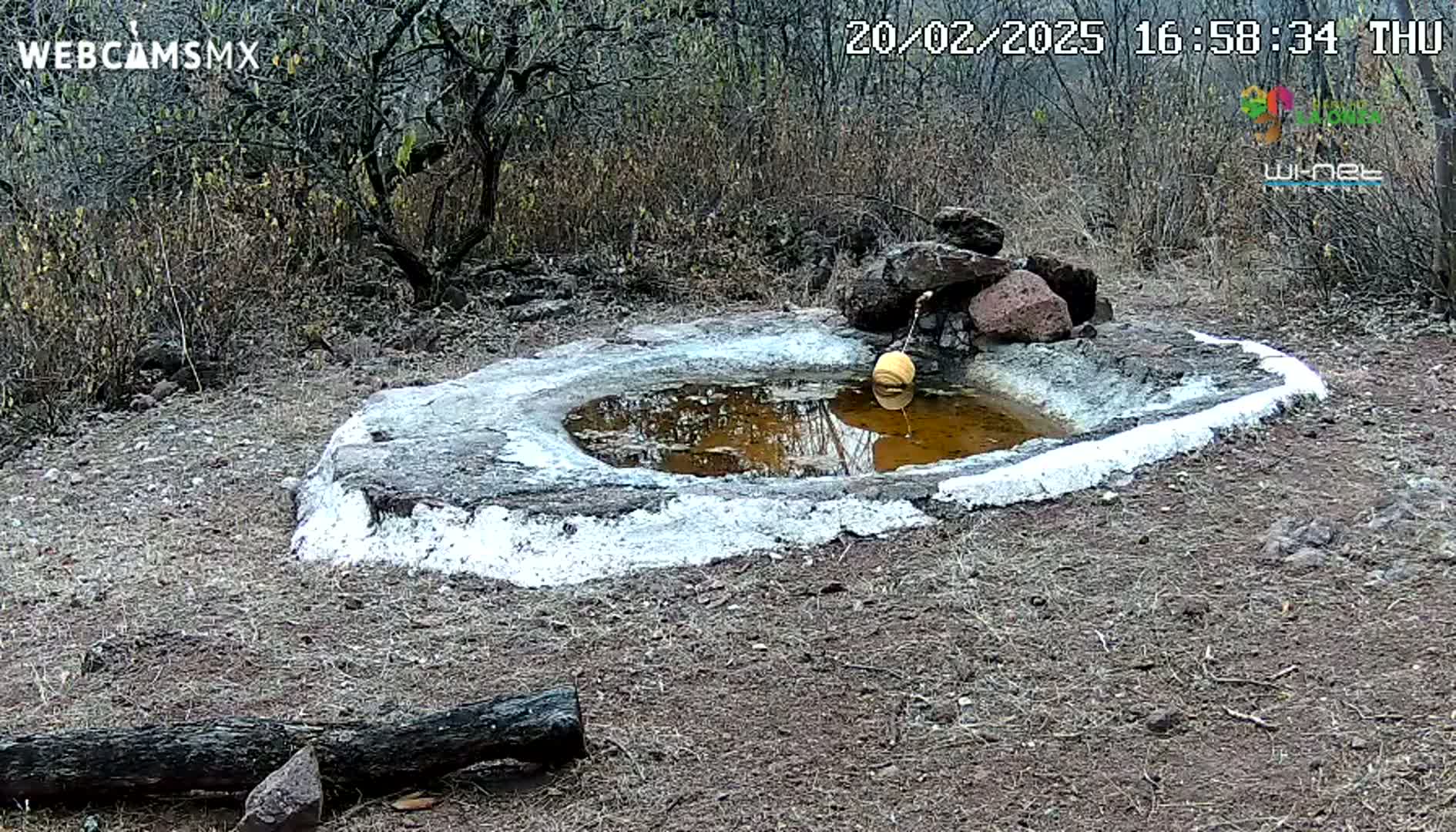 A small, round, stone basin with a shallow pool of muddy water sits in dry, scrubby terrain under a partly cloudy sky.