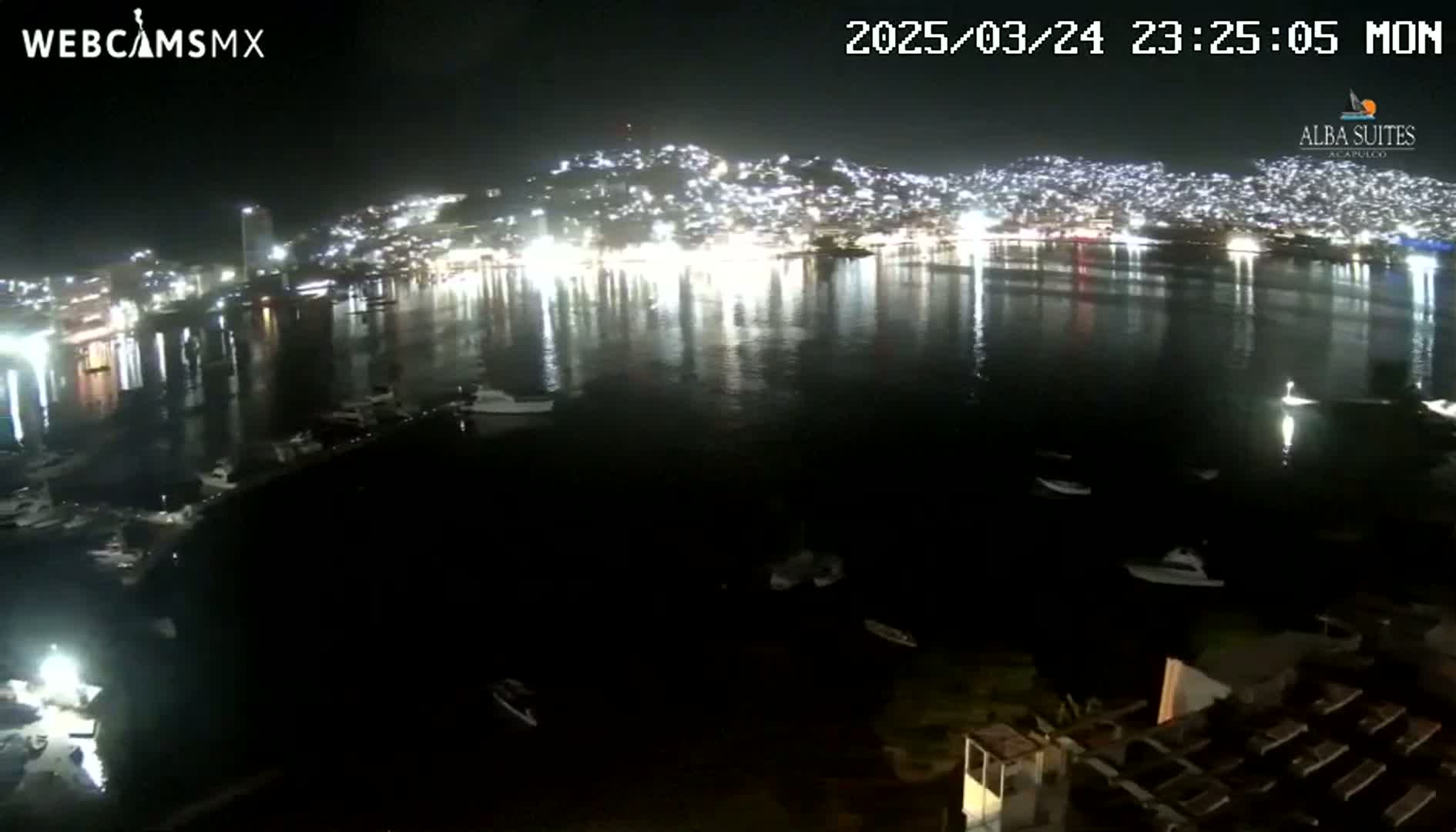 A nighttime view of a city's lights reflected in a calm body of water with several boats moored in the harbor.
