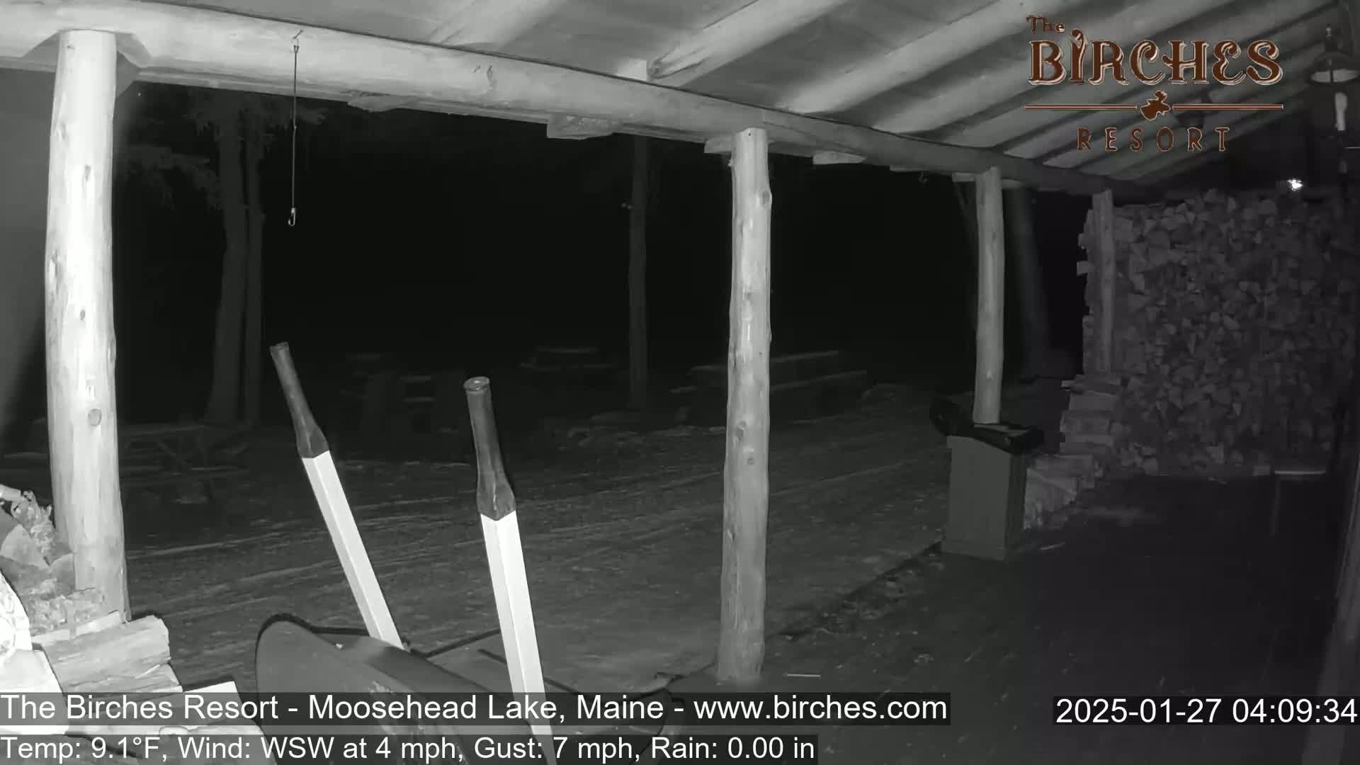 A nighttime, black-and-white image shows a covered porch with a woodpile and a wheelbarrow under rainy conditions.