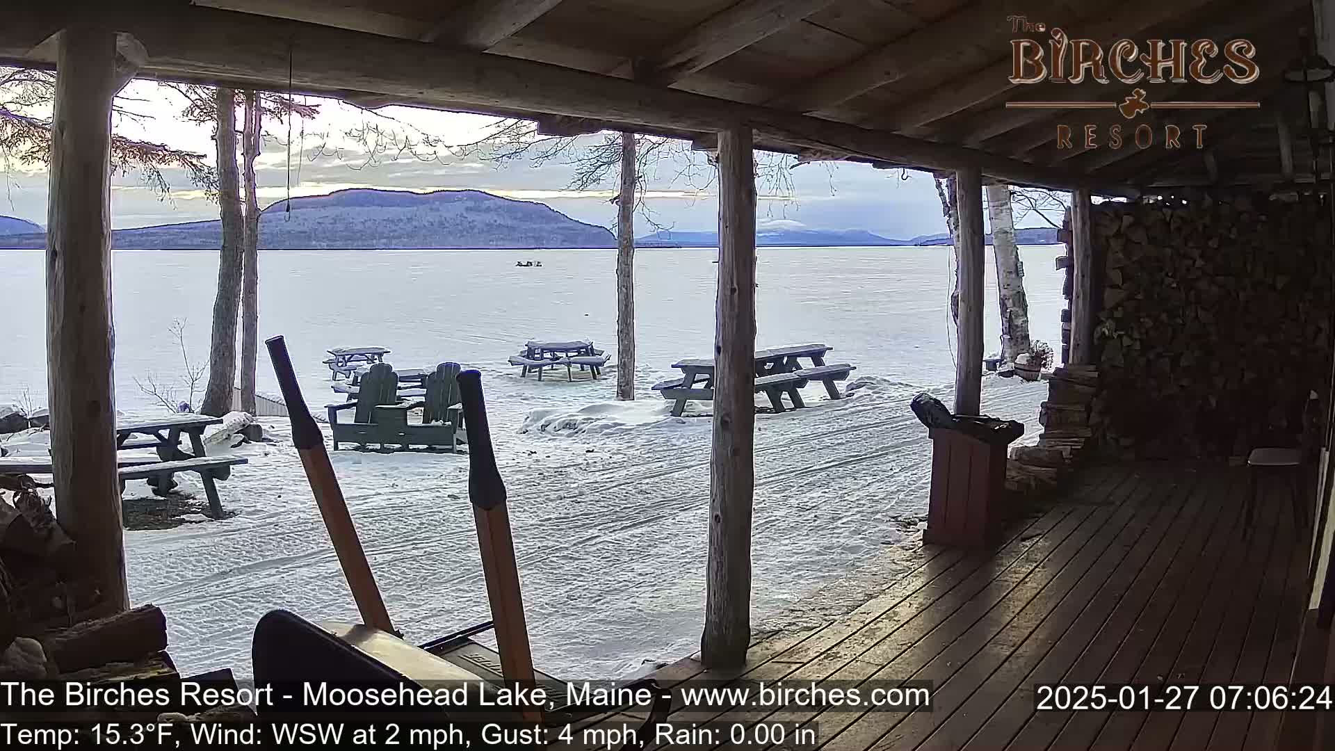 From a snow-covered porch, a frozen lake is visible under a partly cloudy sky, with several picnic tables and chairs scattered on the snow.