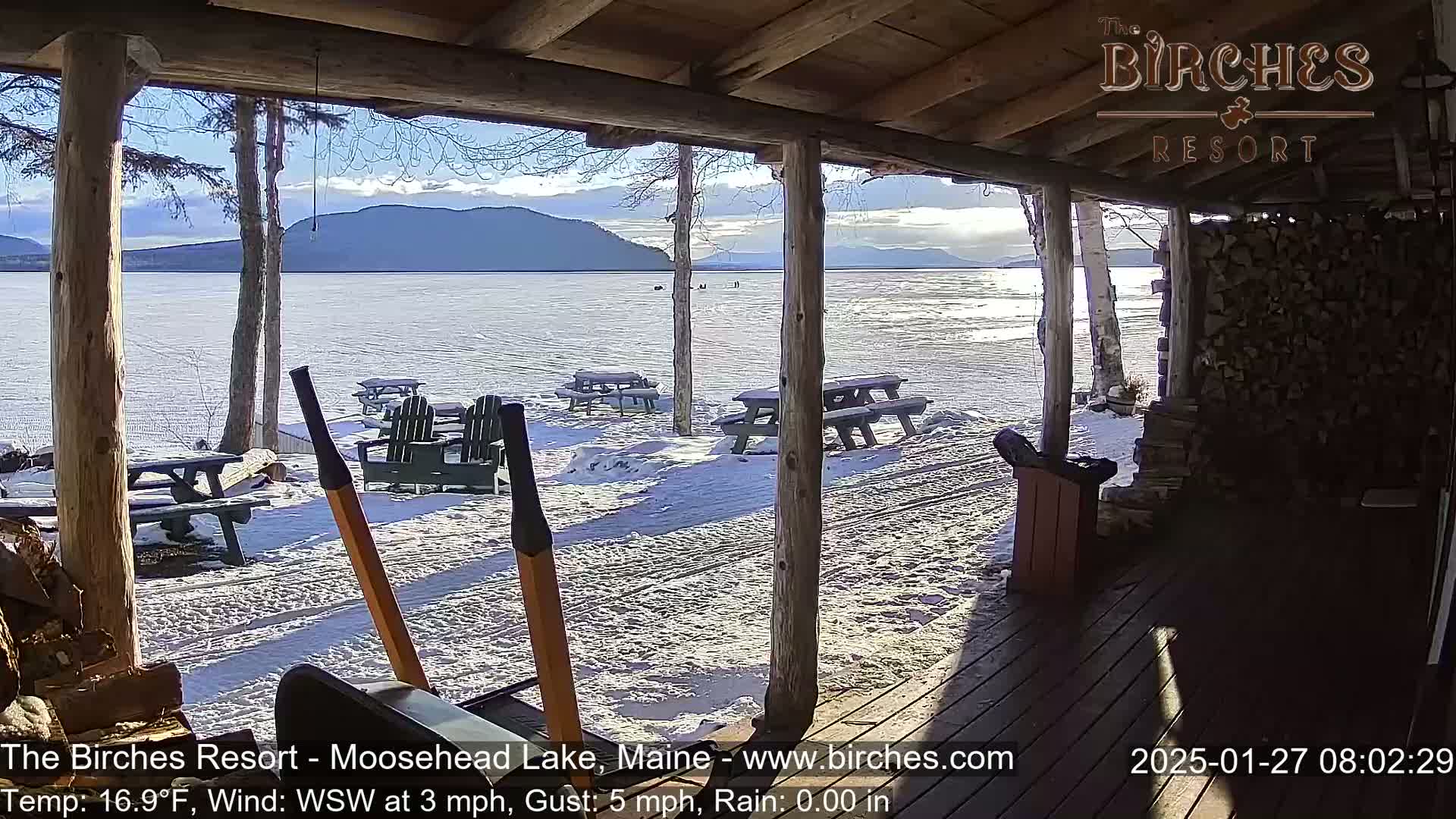 A snow-covered lakeside porch with picnic tables and Adirondack chairs overlooks a partially frozen lake and distant mountains under a partly sunny sky.