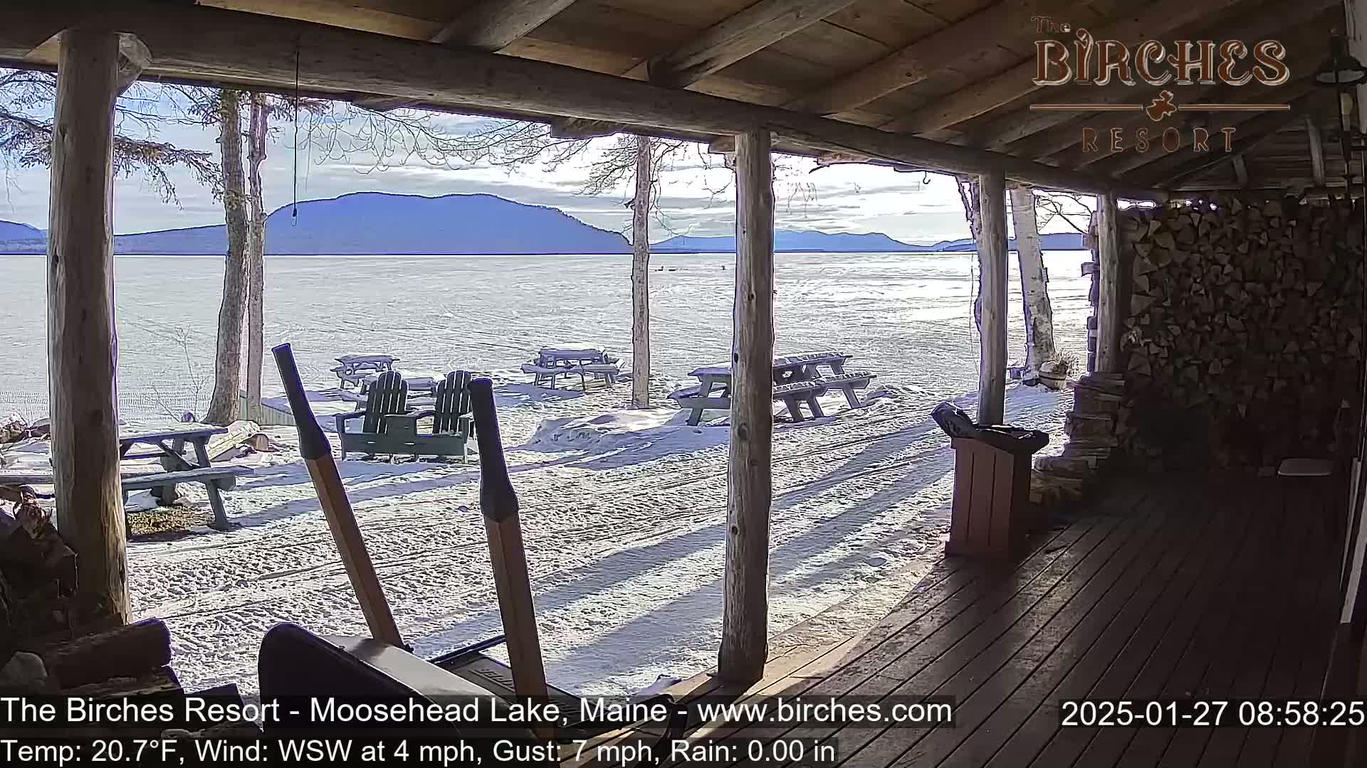 From a covered porch, a sunny winter view shows a snow-covered lakeshore with picnic tables and Adirondack chairs, and a distant mountain range.
