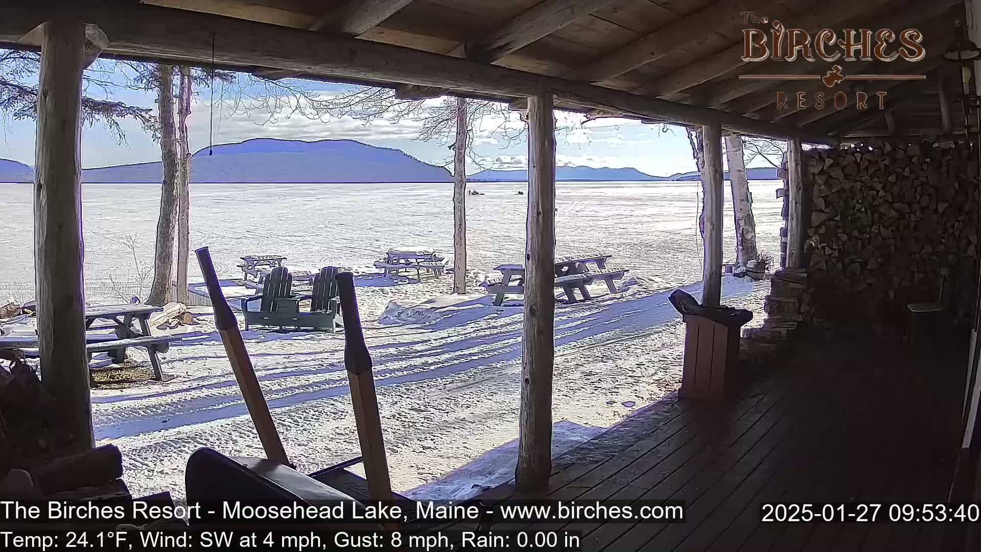 A view from a covered porch shows a snow-covered lake and distant mountains under a sunny sky, with picnic tables and chairs scattered on the snow-covered ground.
