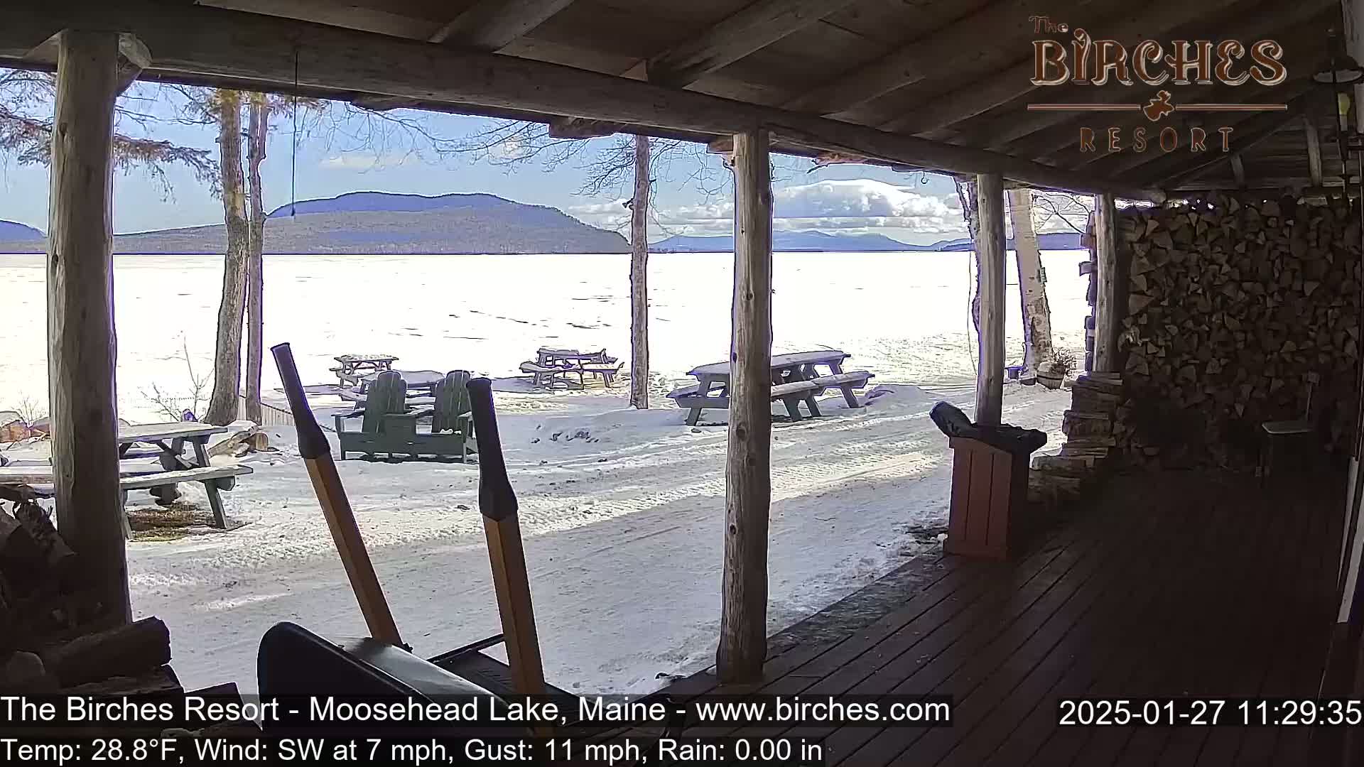 A view from a porch shows a snow-covered lakeside area with picnic tables and a partly sunny sky, and mountains in the background.