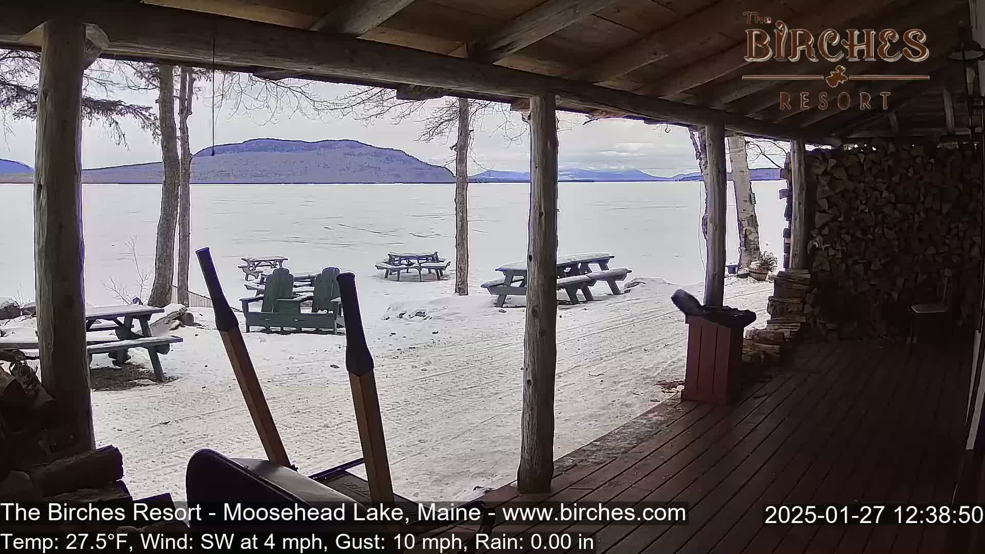 A snow-covered lakeside scene is viewed from a wooden porch, featuring several picnic tables and Adirondack chairs in the snow, with a calm, frozen lake and distant mountains in the background under an overcast sky.