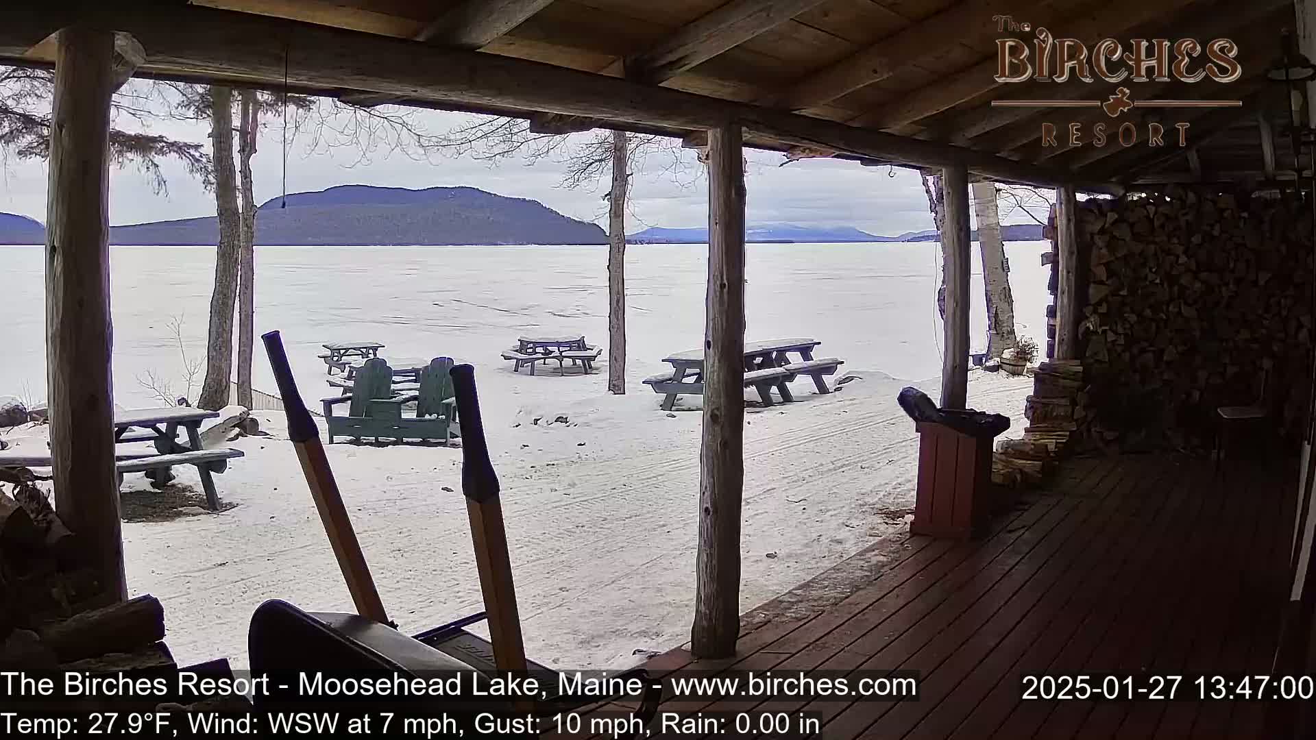 From a snow-covered porch with picnic tables, a view of a frozen lake and distant mountains under an overcast sky is seen.
