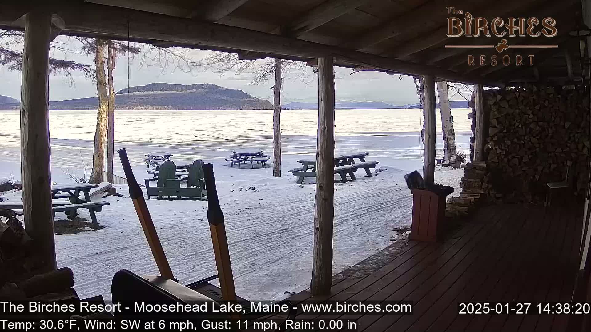 A snow-covered porch overlooks a frozen lake with mountains in the background under a cloudy sky.