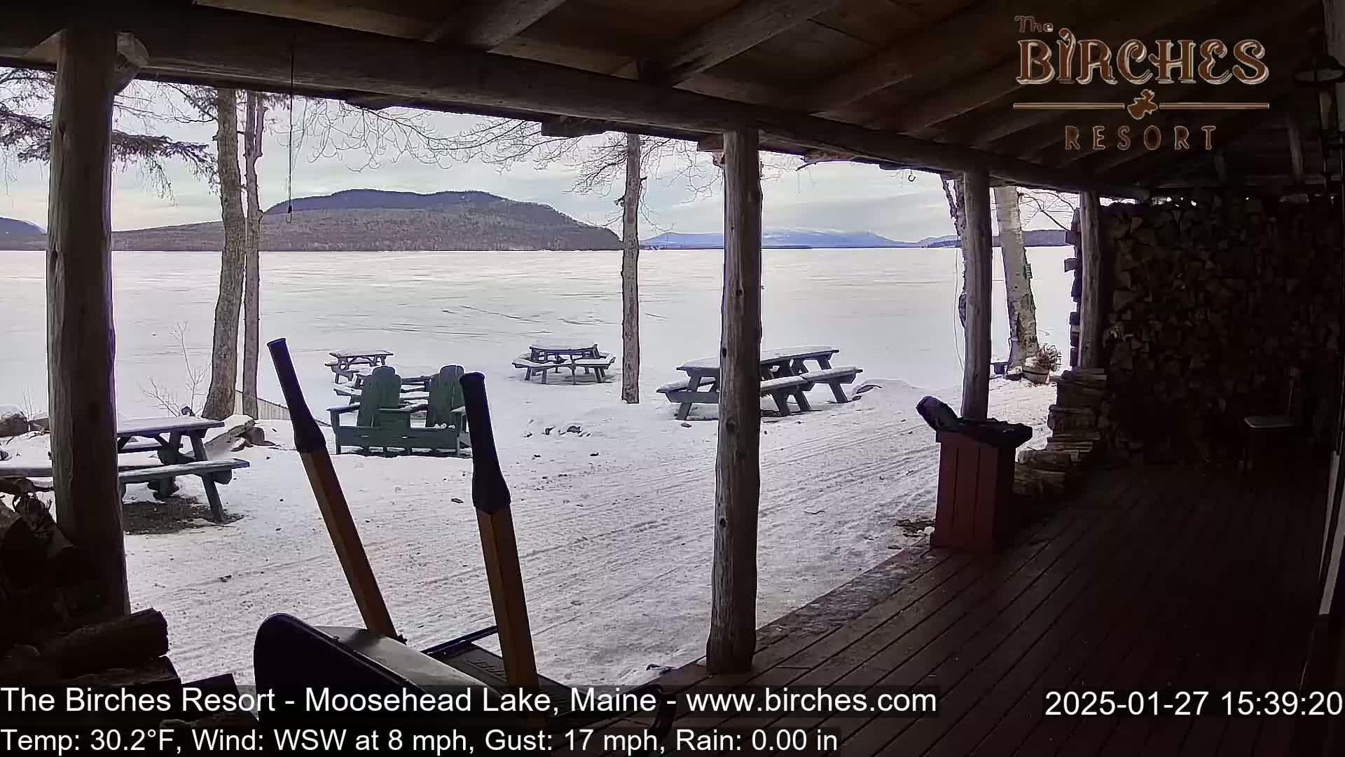 A snow-covered frozen lake is viewed from a wooden porch with picnic tables, a wheelbarrow, and a woodpile in the foreground, under cloudy skies.