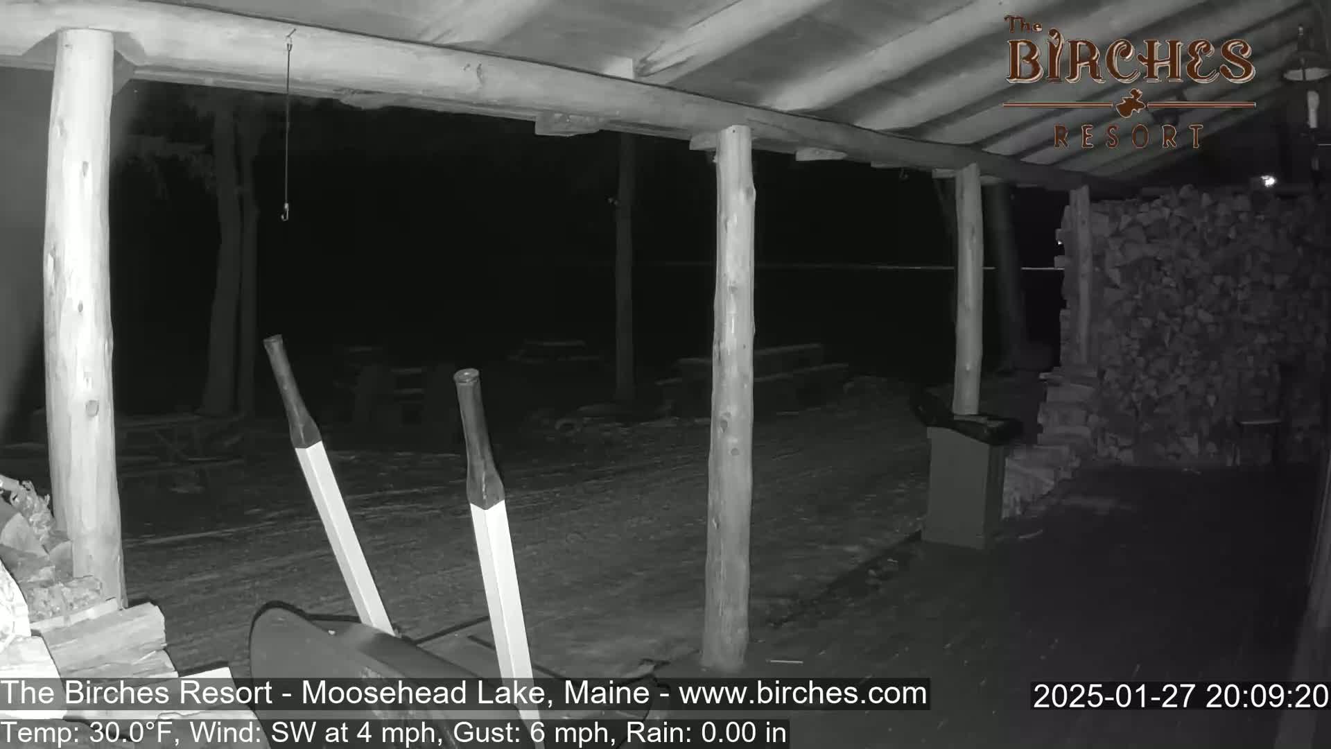 A nighttime, snow-covered outdoor area under a wooden structure, with a large stack of firewood visible, and two handles of what appears to be a snow removal device in the foreground.