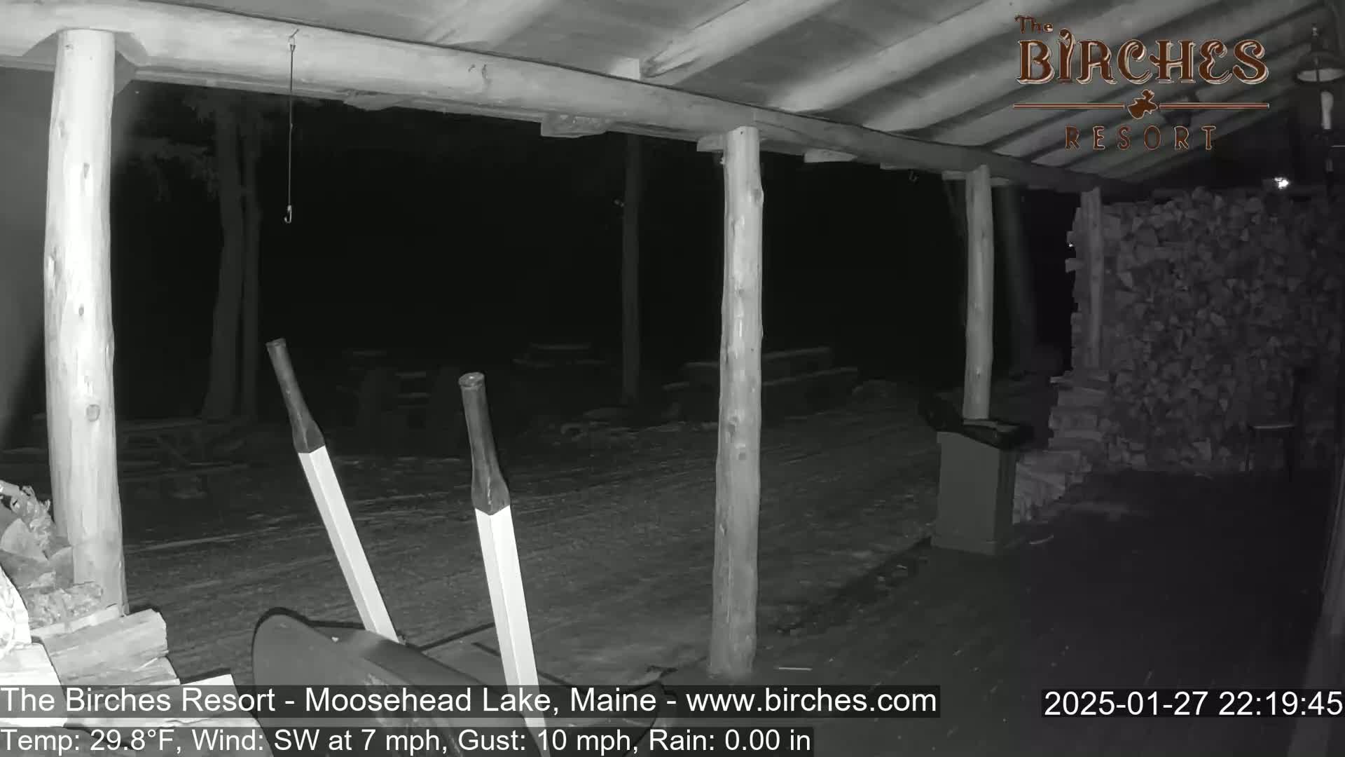 A nighttime, snow-covered outdoor area under a wooden structure with a snow shovel and a pile of firewood visible.