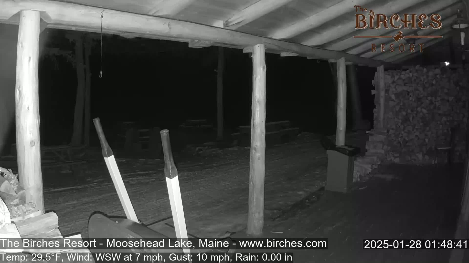 A nighttime, low-light view of an open-air structure with wooden posts and a snow-covered ground, showing a snow shovel and a stack of firewood.