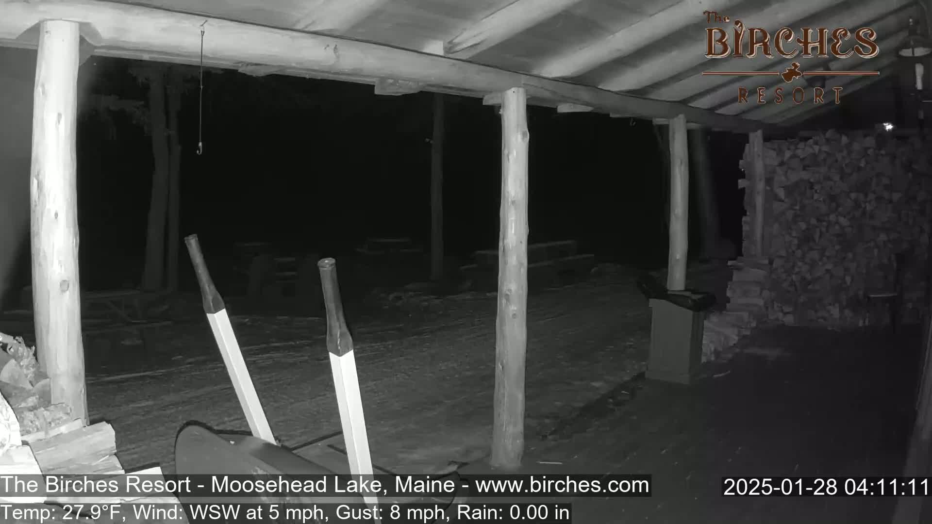 A snow-covered outdoor area at night, partially sheltered by a wooden structure, shows a snow shovel and a large stack of firewood.