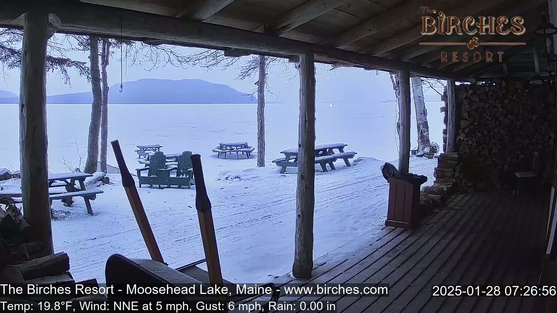 A snow-covered porch overlooks a frozen lake and distant mountains under an overcast sky.