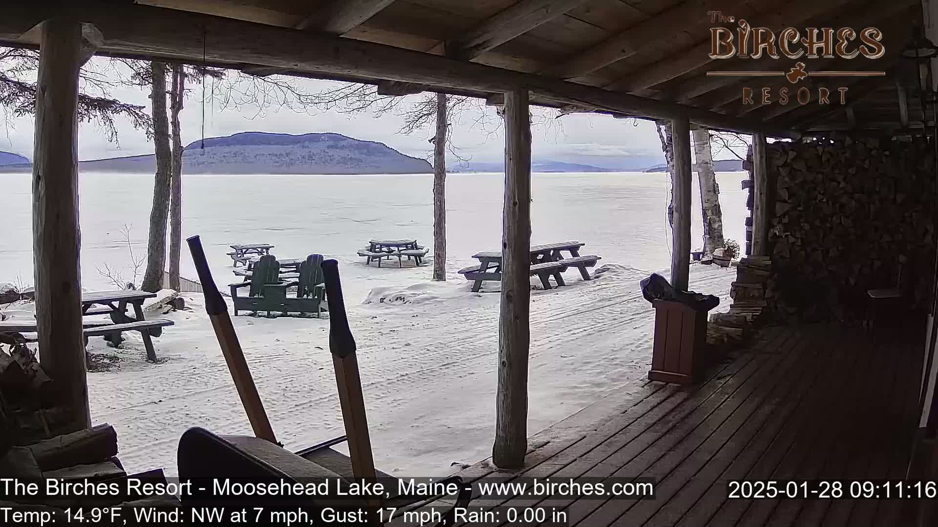 From a snow-covered porch overlooking a frozen lake and distant mountains, several picnic tables and chairs sit empty under a cloudy sky with a northwest wind.