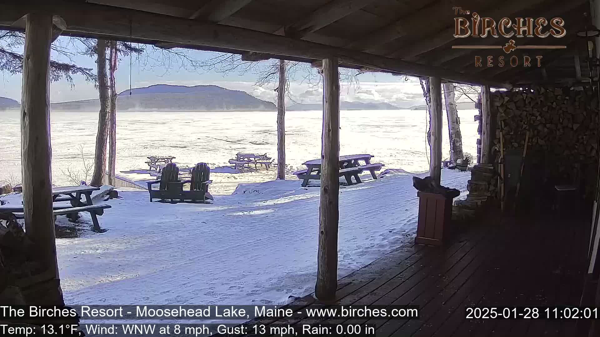 A snow-covered lakeside scene is viewed from a wooden porch, with picnic tables and Adirondack chairs scattered across the snow.