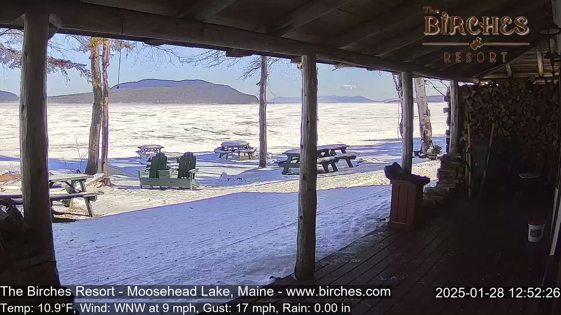 A snow-covered lake and mountains are viewed from a covered porch with picnic tables and Adirondack chairs on a sunny, windy day.