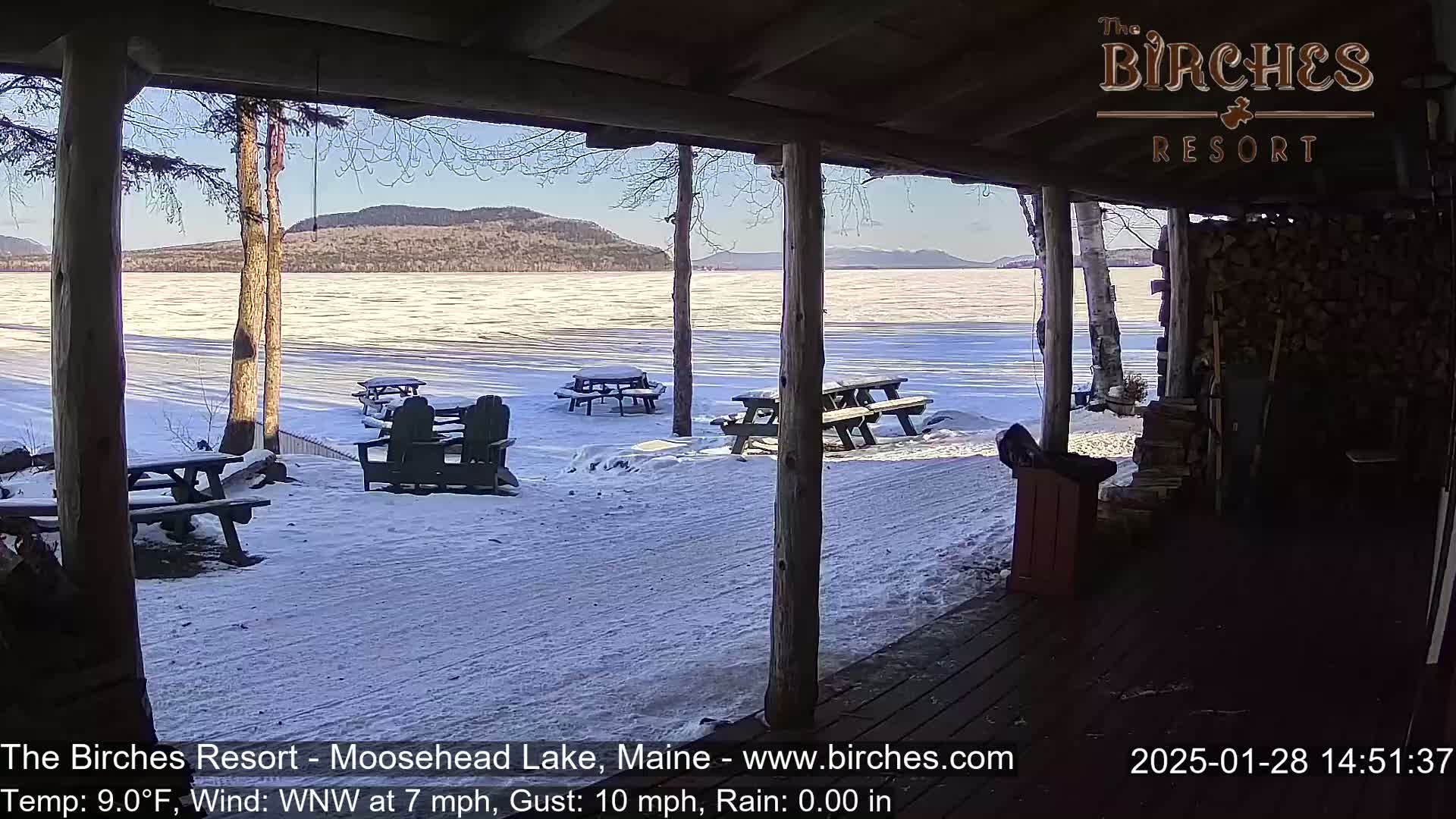 A snow-covered lakeside scene is viewed from a covered porch, featuring picnic tables and Adirondack chairs, with a sunny, cold day and mountains in the background.