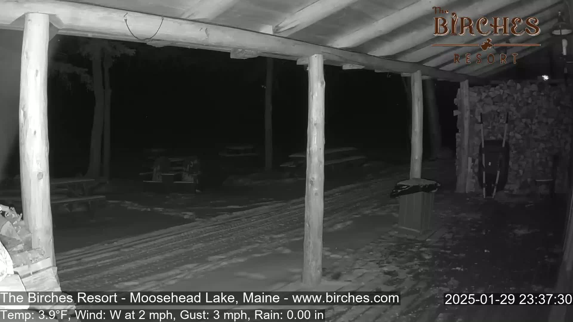 A nighttime, snow-covered outdoor area is seen from under a wooden covered porch, with picnic tables and a woodpile visible in the distance.
