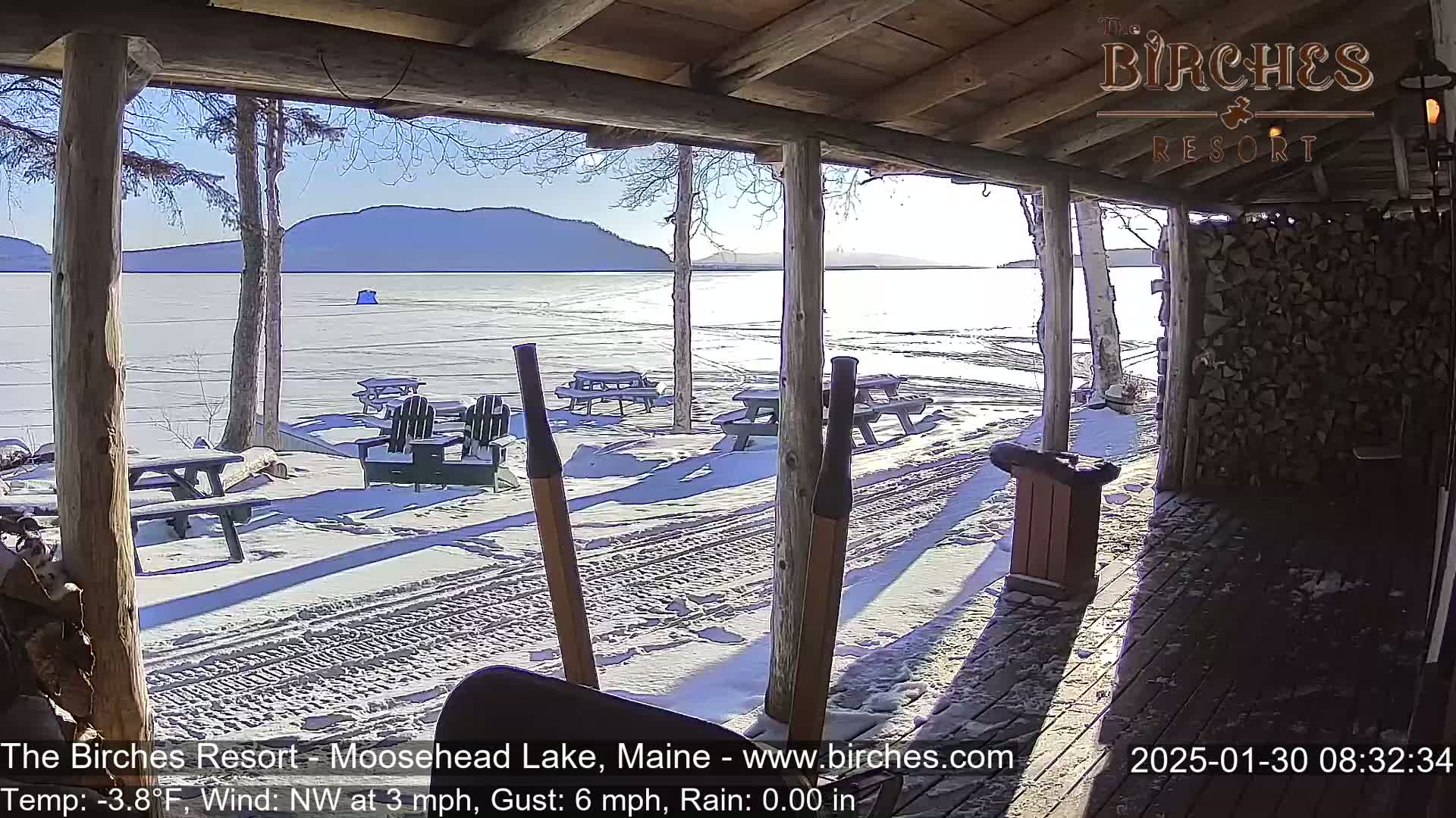 A snow-covered porch overlooks a frozen lake and distant mountains under a sunny sky.