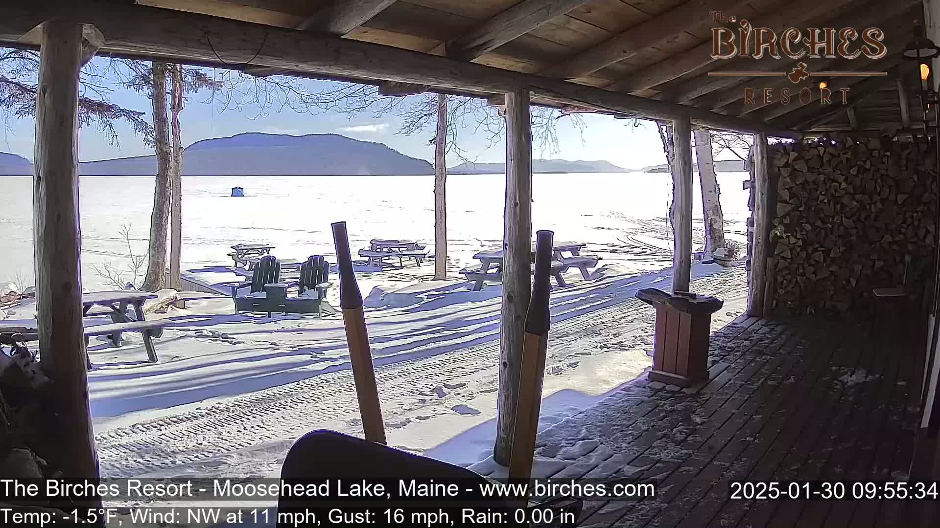 A snow-covered porch overlooks a partially frozen lake and distant mountains under a sunny sky.