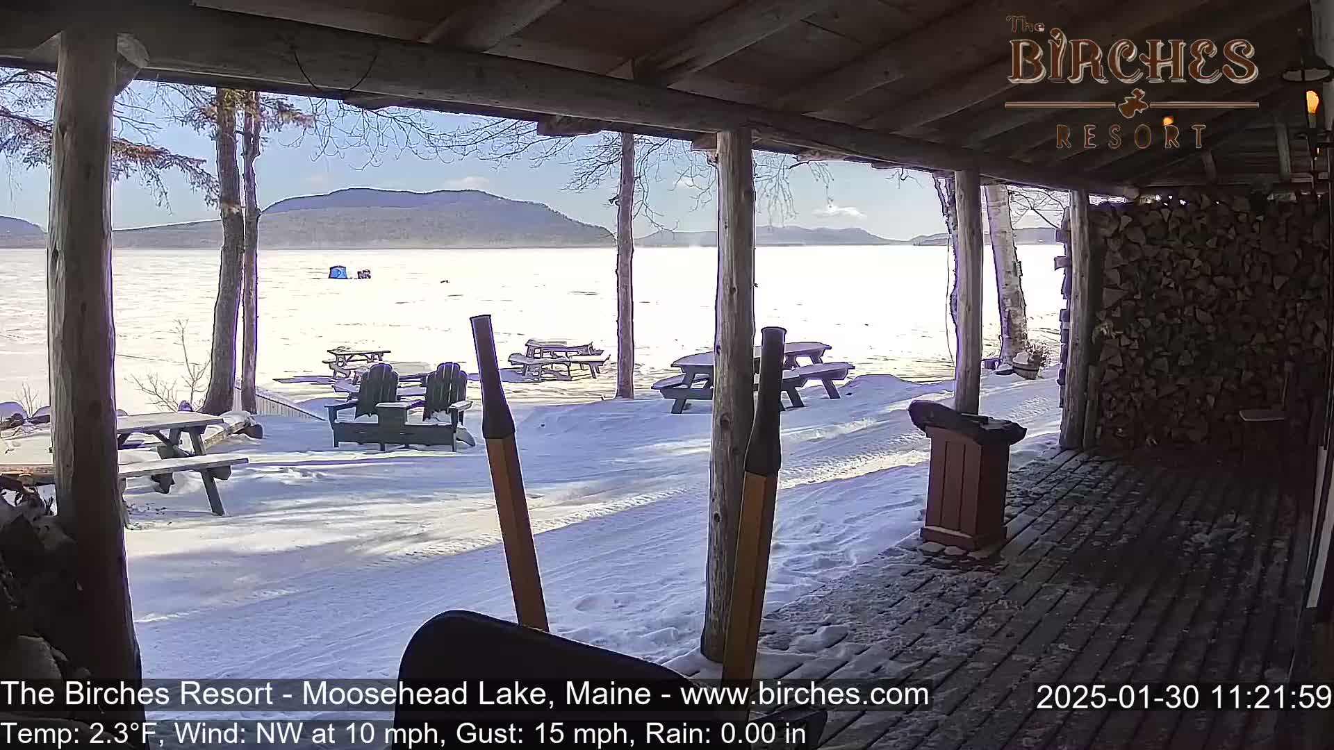 A sunny, snowy view of a frozen lake with picnic tables and Adirondack chairs, seen from a covered porch with a woodpile nearby.