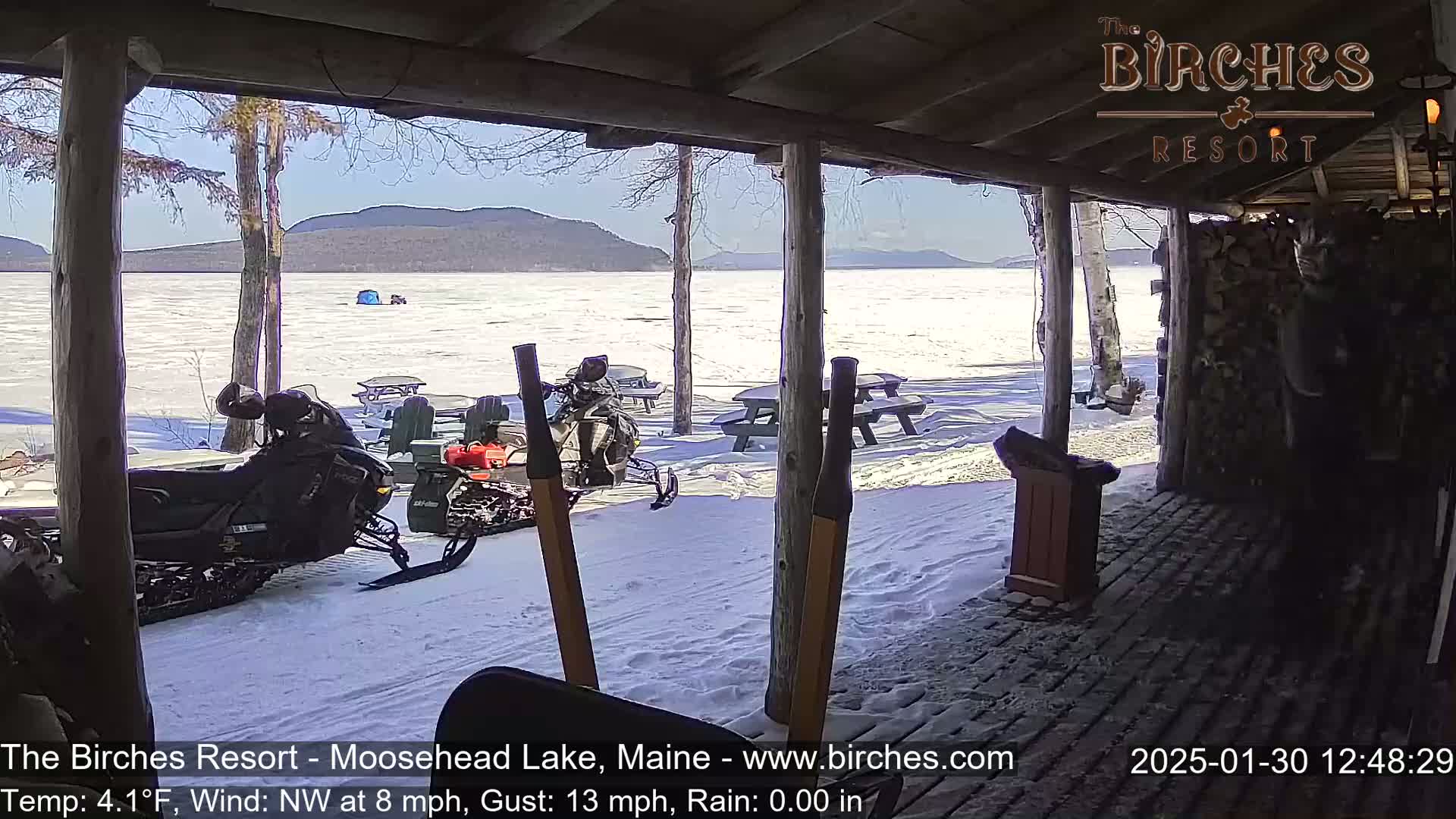 From a snow-covered porch overlooking a frozen lake and distant mountains, snowmobiles are parked near picnic tables under a sunny sky.