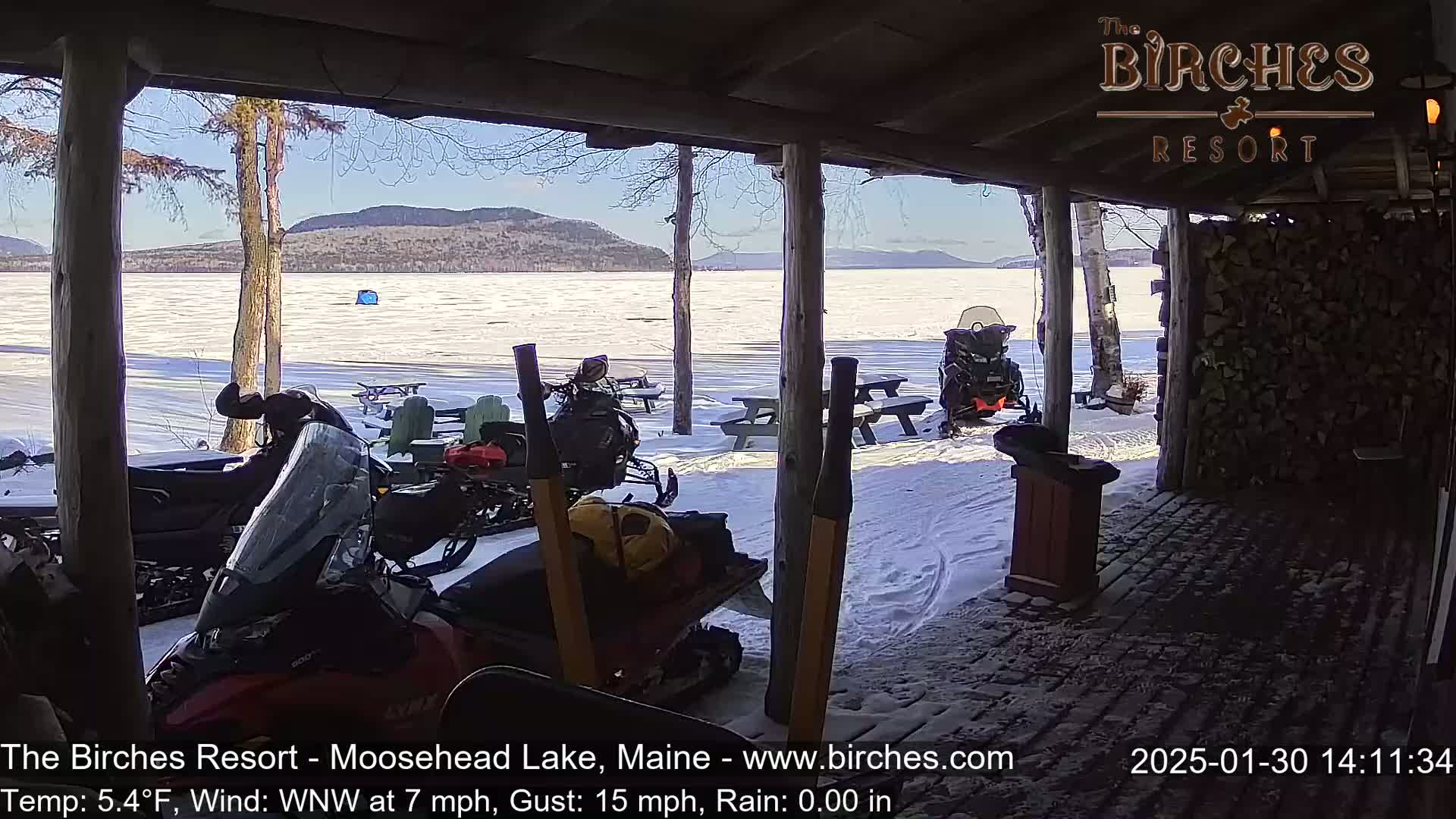 A sunny view of a snow-covered frozen lake and mountains from a covered porch, with snowmobiles and picnic tables visible in the foreground.