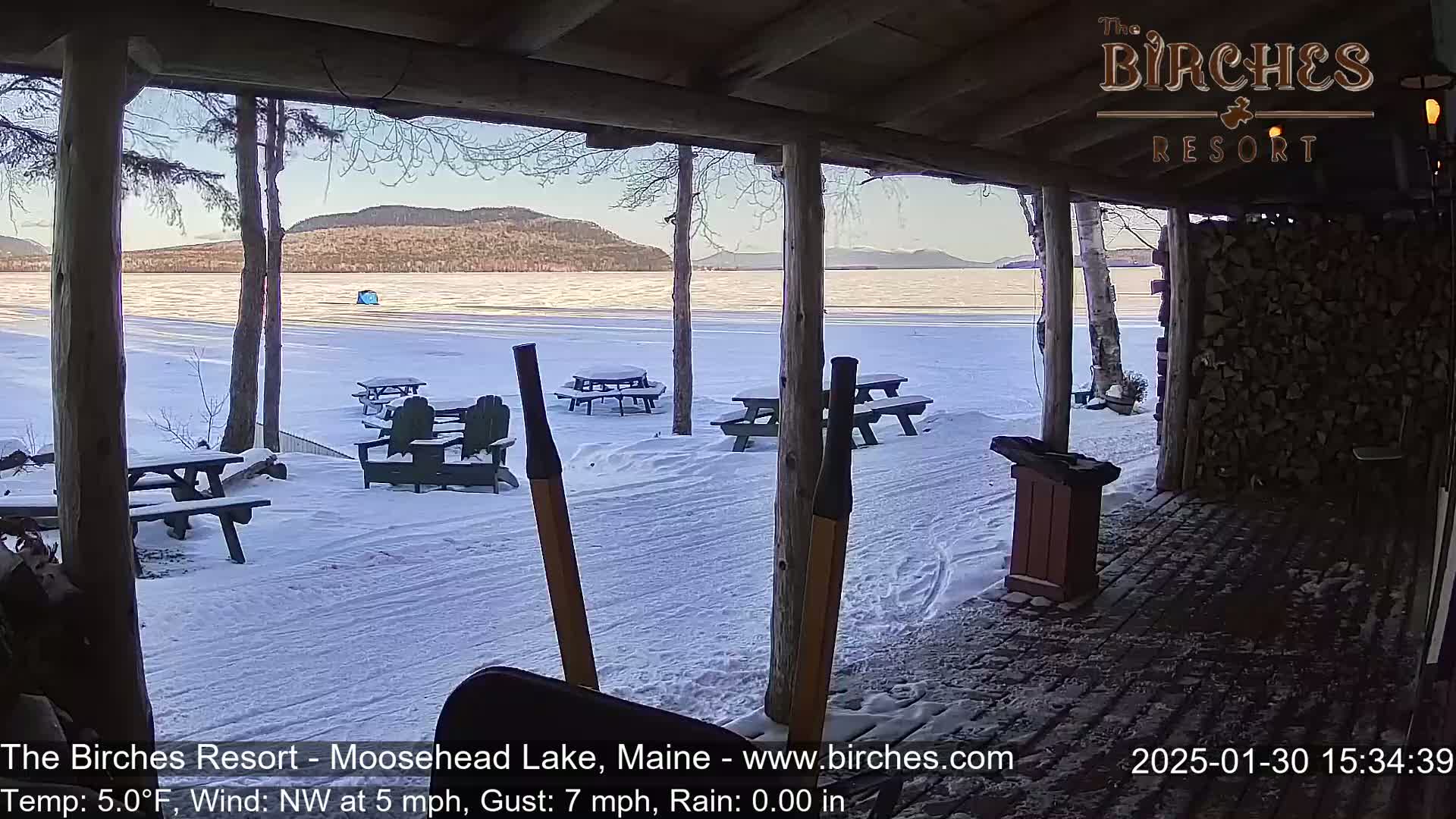 A snow-covered lakeside patio with picnic tables and Adirondack chairs overlooks a frozen lake and distant mountains under a partly sunny sky.