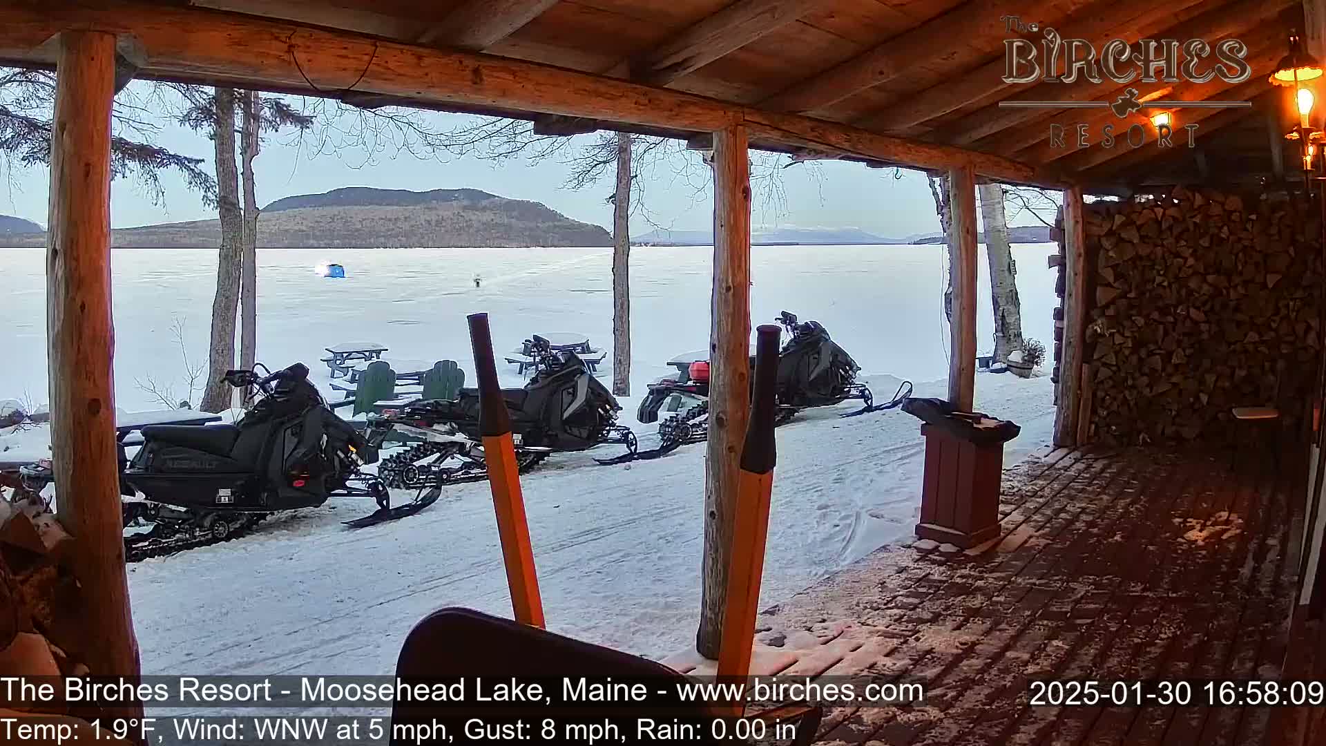 From a snow-covered porch overlooking a frozen lake and distant mountains, two snowmobiles are parked near outdoor seating.